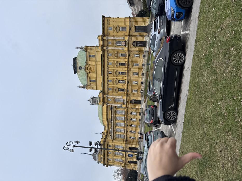 The image displays an exterior view of the Palace of the Croatian Academy of Sciences and Arts, situated at Trg Nikole Šubića Zrinskog 11, City of Zagreb, Croatia. The prominent, multi-story building features a yellow facade with classical architectural details, including decorative columns and multiple green domes, dominating the background under an overcast sky. In the lower-left foreground, a human left hand is positioned, forming a "thumbs down" gesture. The foreground consists of a grassy area with some visible soil patches, separated by a curb from a paved parking area. Multiple vehicles are parked in this area; a black sedan (BMW 7 Series type) is centrally located, with a light grey SUV (Hyundai Ioniq 5 type) partially visible to its left and a blue SUV (Ford Puma type) to its right. A decorative black street lamp, equipped with multiple luminaires and a blue parking sign, stands on the left side of the frame. Park benches are discernible in the distance between the parked cars and the building. The lighting is diffused and uniform, consistent with an overcast day.