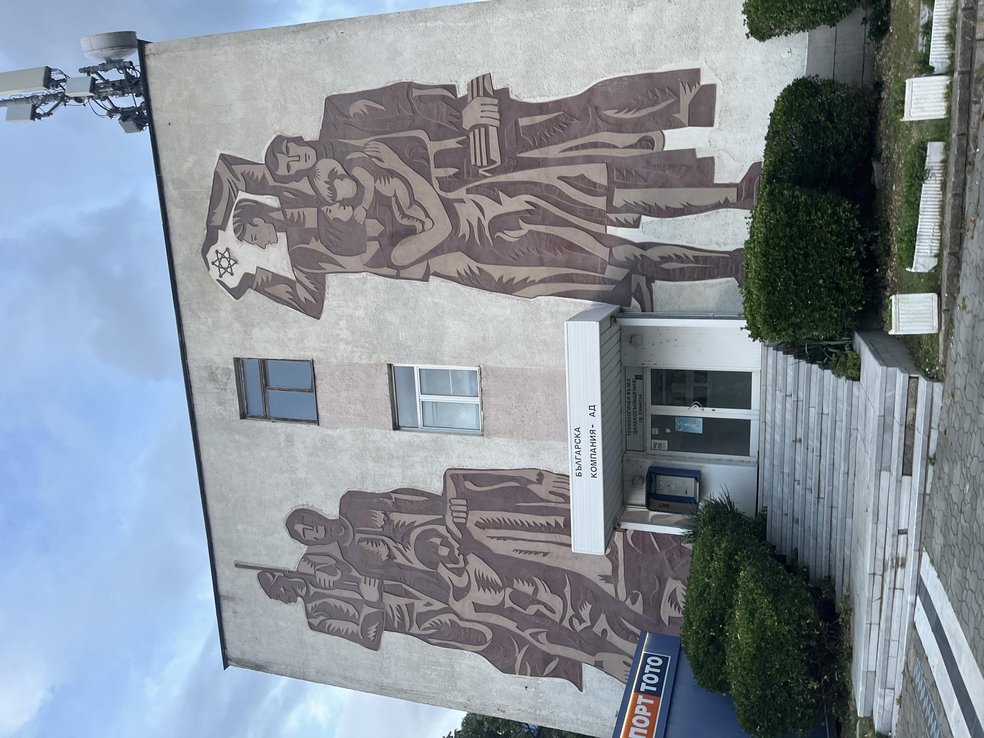 A low-angle shot captures a building in Simitli, Bulgaria, adorned with a large mural. The mural, rendered in shades of brown and cream, features figures in a socialist-realist style. The upper part of the mural depicts a male figure holding a Star of David, with other figures surrounding him, including what appears to be a mother holding a child. The lower part of the mural depicts figures holding weapons. Two windows punctuate the wall between the upper and lower portions of the mural.
To the right of the mural is the entrance to the building, marked by a white canopy that reads "Българска Компания - АД" and "Технологичен възел далекосъобщения Гр. Симитли". A blue sign that reads "Порт Тото" is visible near the bottom of the frame.
Green bushes grow in front of the building's entrance. To the right, a set of stairs lead to the entrance, and grey cobblestones are visible on the ground. In the upper left corner of the image, a telecommunications tower extends from the building, contrasting with the classic mural. The sky is cloudy.