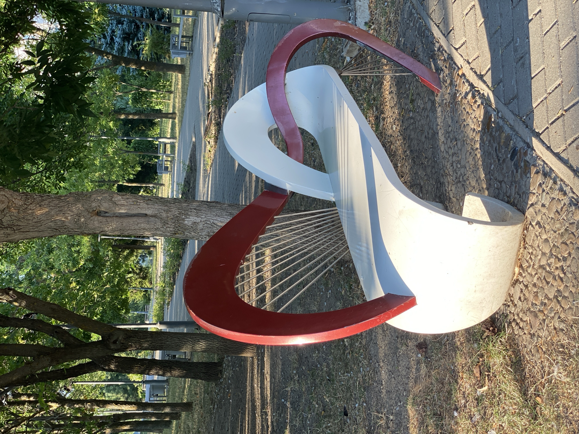 The image shows a uniquely designed park bench in the shape of two intertwined hearts, one red and one white, located on a cobblestone and paved walkway in Chișinău, Moldova. The bench is situated outdoors, surrounded by trees and grass, with a path extending into the background. The lighting suggests it is daytime, with sunlight casting shadows on the ground. There are no people visible in the image.