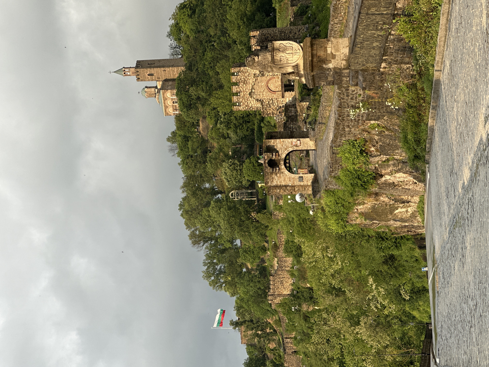 The image captures a side view of Tsarevets Fortress, a historic stronghold in Veliko Tarnovo, Bulgaria. The fortress complex is perched atop a hill covered with lush greenery. Stone walls and structures of varying heights, indicative of different construction periods, are visible along the hillside. A Bulgarian flag is flying near the bottom of the hill. Higher up, a distinct stone tower and a church with a dome are seen, representing the core of the fortress's religious and defensive architecture. The sky is overcast and grey, providing a muted backdrop to the scene. A wet road surface occupies the foreground, suggesting recent rainfall. No people are visible in the shot.