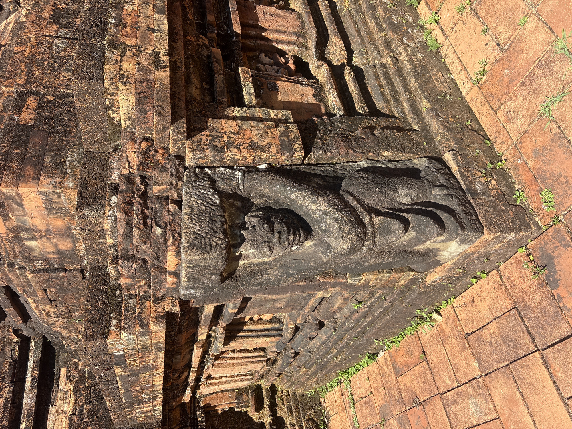 The image depicts a corner of an ancient brick structure, likely a temple or ruin, located in Đà Nẵng, Vietnam. A stone carving of a grotesque or guardian figure is integrated into the corner, supporting what appears to be a lintel or decorative element. The structure is made of weathered red bricks, some of which are covered in dark moss or lichen. The architectural style suggests a historical Asian influence. The foreground is paved with terracotta-colored bricks, with small green plants growing in the crevices between them and around the base of the structure. The lighting indicates a bright, sunny day, casting shadows on the textured surfaces of the brickwork and the stone carving. The scene is devoid of people, suggesting a quiet, historical site.