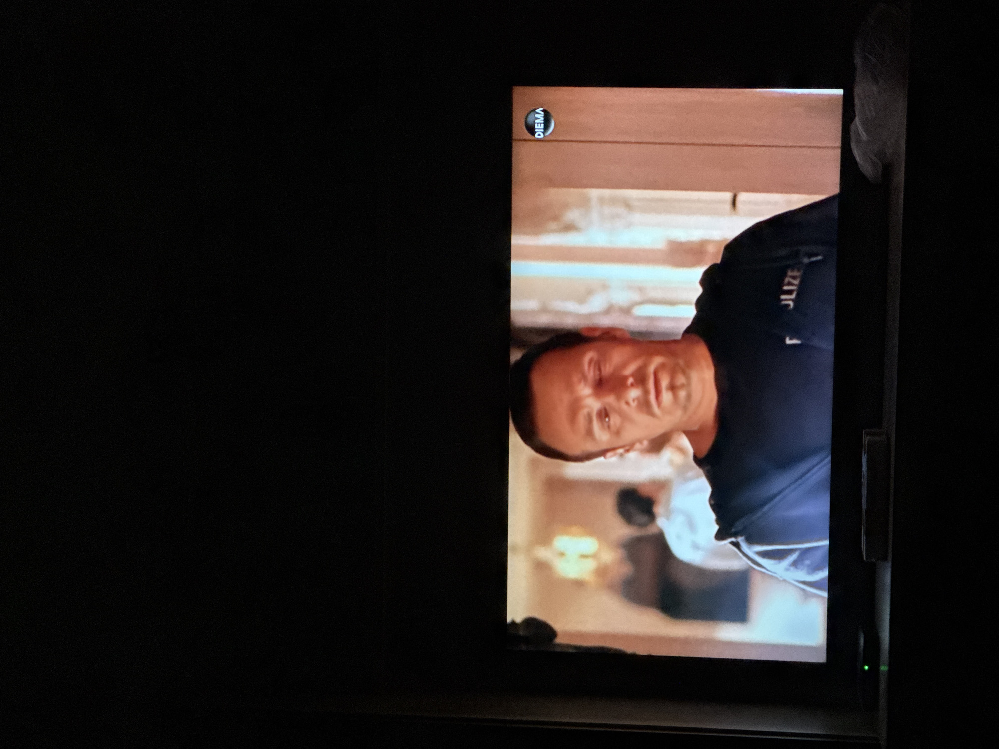 A screen displays an image of a man wearing a dark blue or black shirt with "POLIZEI" printed on it. The image is slightly tilted. The "DIEMA" logo is visible in the upper left corner of the screen image. A person partially visible in the background is wearing a white garment. The TV or screen is in a dark room. I can't be certain that this is Sarnevo, Bulgaria because no details or objects point to it.