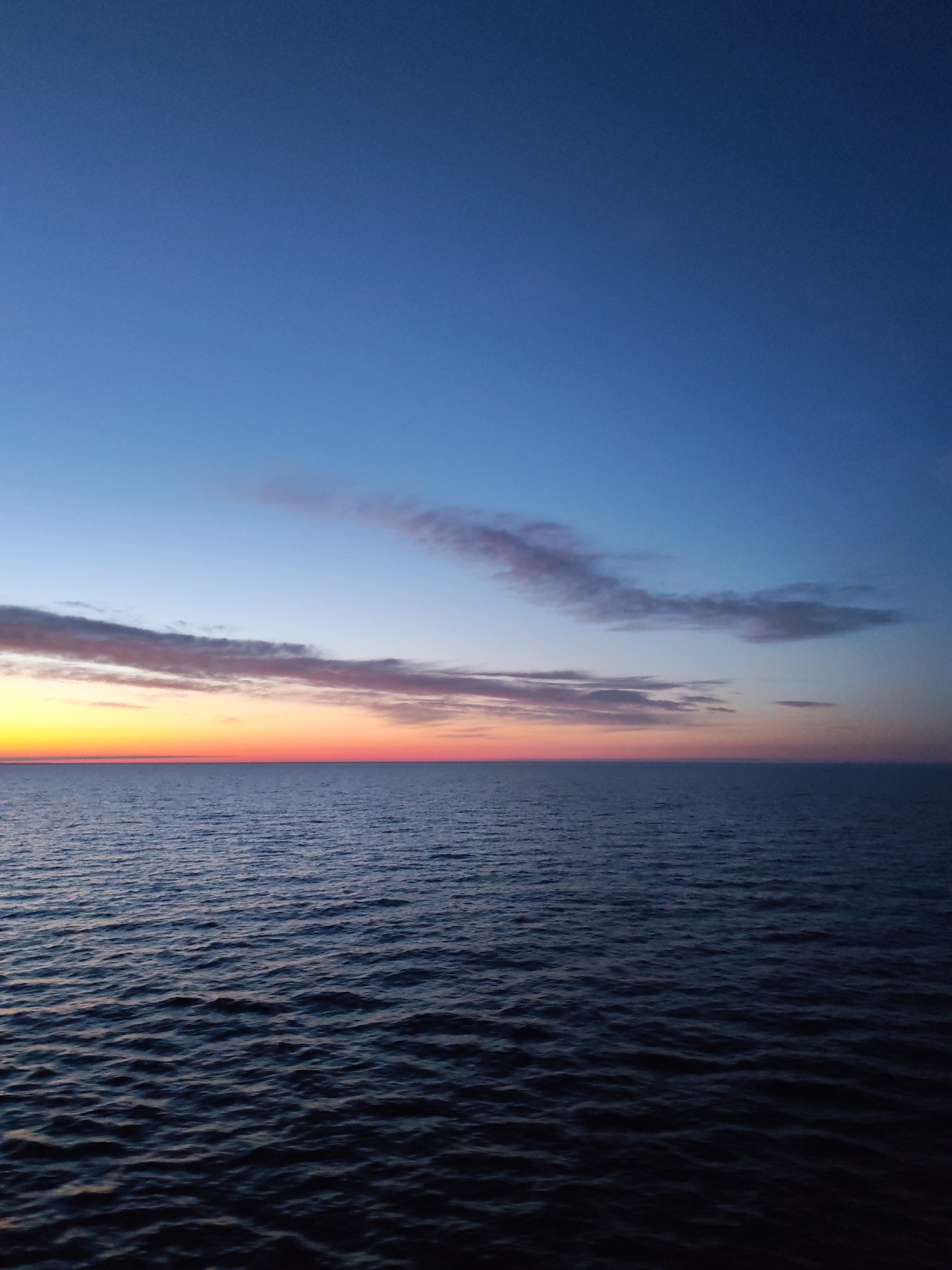 The image shows a twilight scene over water, likely a sea or large lake. The sky transitions from a deep blue at the top to a band of orange and yellow near the horizon, indicating either sunrise or sunset. Several streaks of clouds are present, tinted with pink and gray hues. The water is dark and slightly choppy, reflecting the colors of the sky. The location, Unknown, Russia, is inferred from the prompt, though not evident in the image itself. There are no people or man-made objects visible in the scene. The dominant features are the natural elements: the sky, clouds, and water.