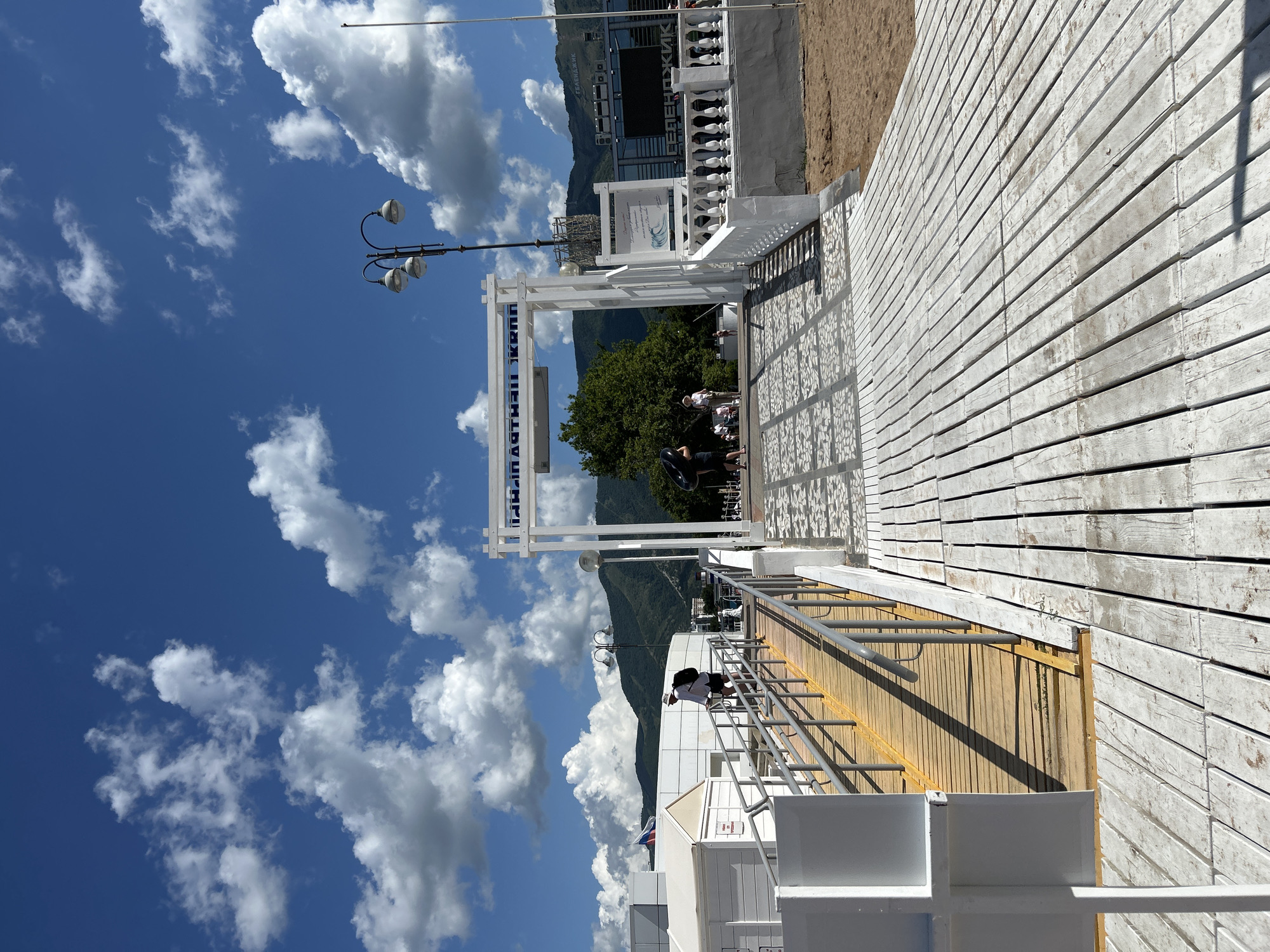The image depicts a sunny day on a boardwalk in Gelendzhik, Russia. The boardwalk, constructed of white-painted wooden planks, leads towards a white archway with Cyrillic text that reads "ПЛАСТИНА КОНА" which translates to "Plastic Cone". Beyond the archway, a staircase with white balustrades ascends. In the background, lush green mountains rise against a bright blue sky filled with fluffy white clouds. The scene is populated by a few individuals. To the left, a person wearing a white hat and backpack walks up a ramp. Further in the background, several people are gathered near the archway and the staircase. The overall atmosphere suggests a recreational area, likely a beach or promenade, given the sand visible in the lower right portion of the frame.