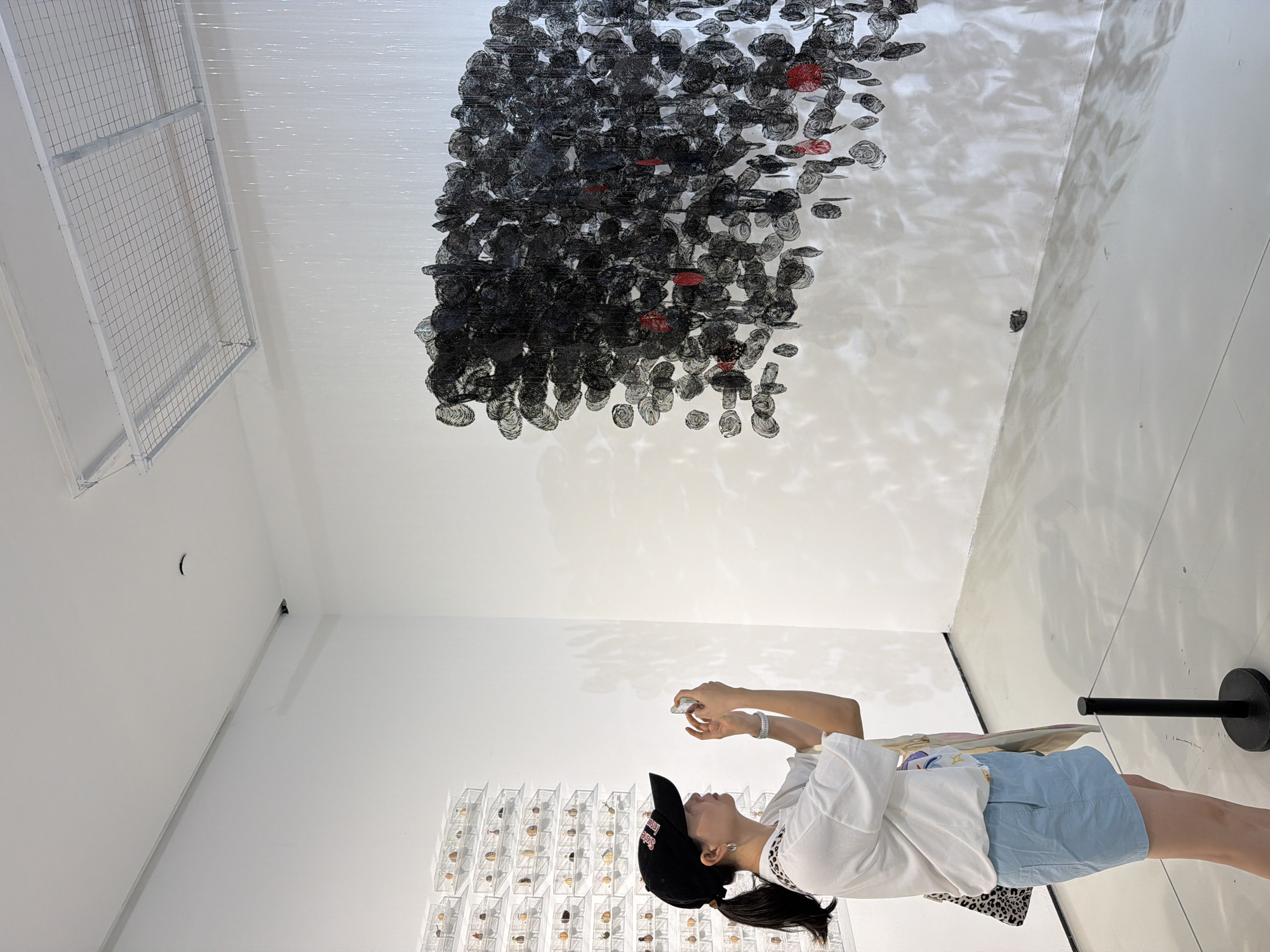 The image depicts an indoor scene within an art gallery or exhibition space in the Chongqing High-tech Zone, China. A woman, positioned in the left foreground and facing right in profile, is actively photographing artwork with a light-colored smartphone held in both hands. She wears a black baseball cap with red and white text, a white short-sleeved t-shirt, and light blue shorts. Her dark hair is in a low ponytail, and she has an earring in her visible left ear and a white wristband. A leopard-print shoulder bag and a light-colored tote bag are carried on her left side.
On the right wall, a large art installation features a dense, cloud-like arrangement of numerous small, black, swirling or circular elements, interspersed with several prominent red circular forms. Above this installation, a white metal grid structure is suspended from the ceiling, from which numerous translucent or white vertical strands descend, creating a visual effect resembling falling rain. Shadows from this artwork are cast onto the white wall and the light gray concrete floor.
On the left wall, further back, another artwork consists of multiple rows and columns of small, clear, rectangular display boxes, each containing a distinct, small object. The walls of the space are uniformly white, and a thin, silver-colored metal track runs horizontally along the upper section of the left and corner walls. A black cylindrical stanchion with a round base stands on the floor in the lower foreground.