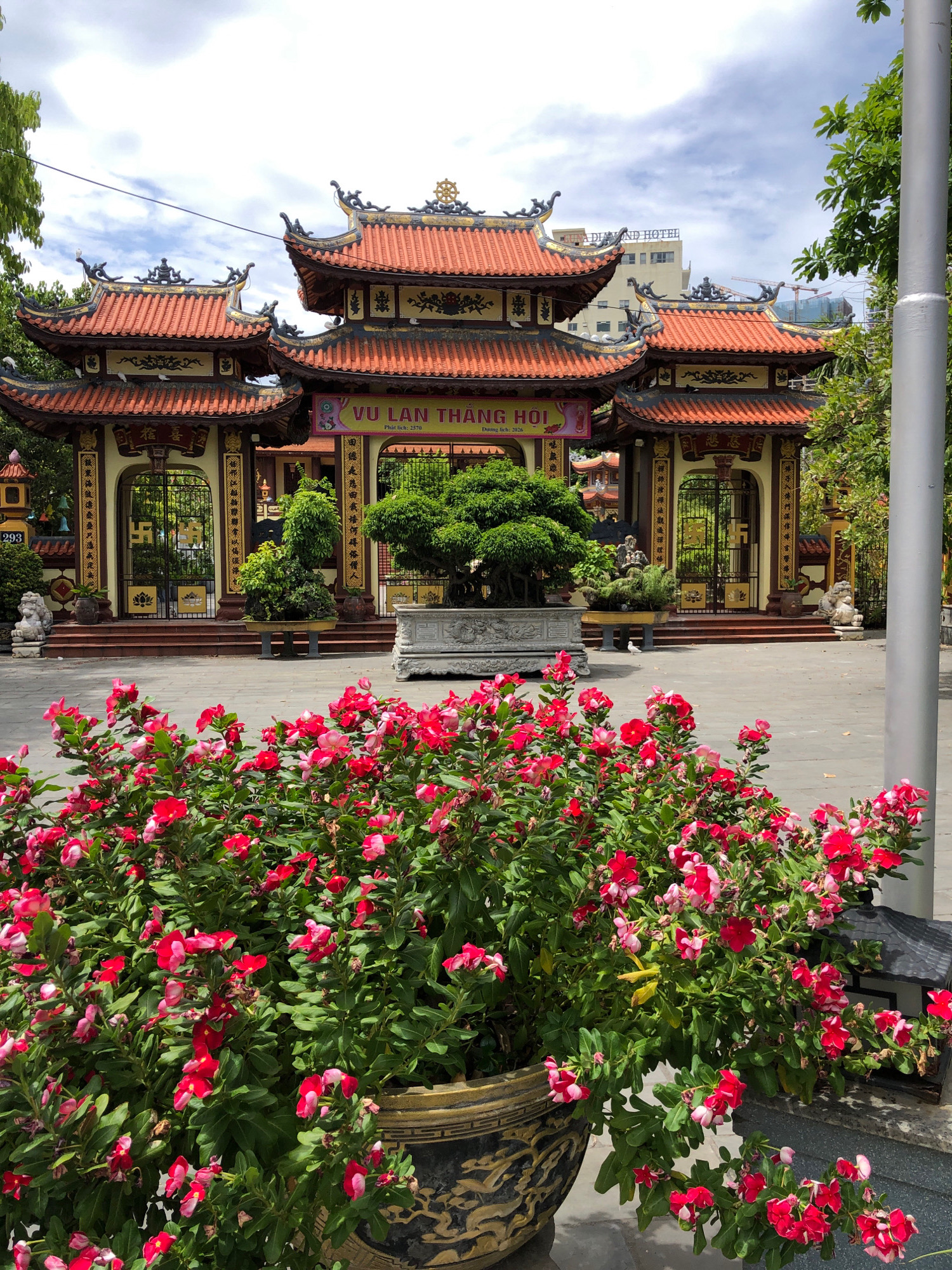 The image depicts a vibrant courtyard in front of a Buddhist temple in Da Nang, Vietnam. In the foreground, a large planter overflows with lush green foliage and numerous bright red and pink flowers. Behind the flowers, a paved courtyard leads to the temple entrance. The temple features traditional Vietnamese architecture with ornate, multi-tiered red roofs, intricate carvings, and yellow accents. Two grand entrances, flanked by decorative gates and guardian lion statues, are visible. A banner with Vietnamese script hangs above the central entrance. In the background, a modern hotel building stands taller than the temple, suggesting an urban setting. The sky is partly cloudy. A tall, slender gray pole stands to the right of the frame.