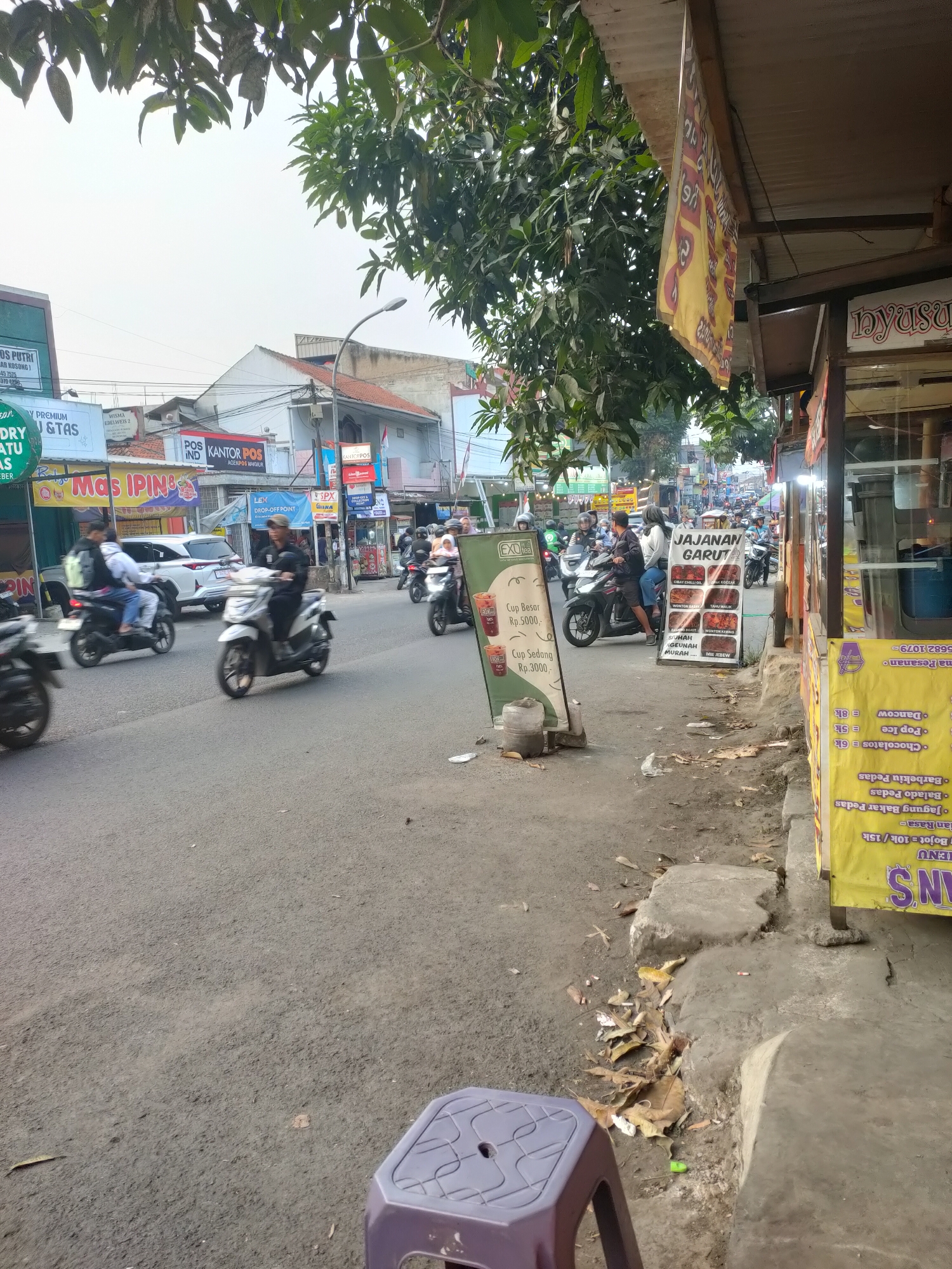 The image captures a street scene in Cimahi, Indonesia, likely a commercial area with several shops and eateries. In the foreground, a grey asphalt road is visible, with a purple plastic stool placed on the side. Several motorcycles are in motion, some occupied by individuals. On the right side of the road, a food stall is set up with a yellow banner displaying a menu in Indonesian, featuring items like "Jajanan Garut," "Wonton Bakso," and "Mie Jeebow." A large tree with green leaves partially obscures the view of the sky and buildings in the background. The buildings are of various designs and heights, some appearing to be shops or residences. Signs for "Kantor POS" (Post Office) are visible on several buildings. The overall atmosphere suggests a bustling urban environment, likely during the daytime.