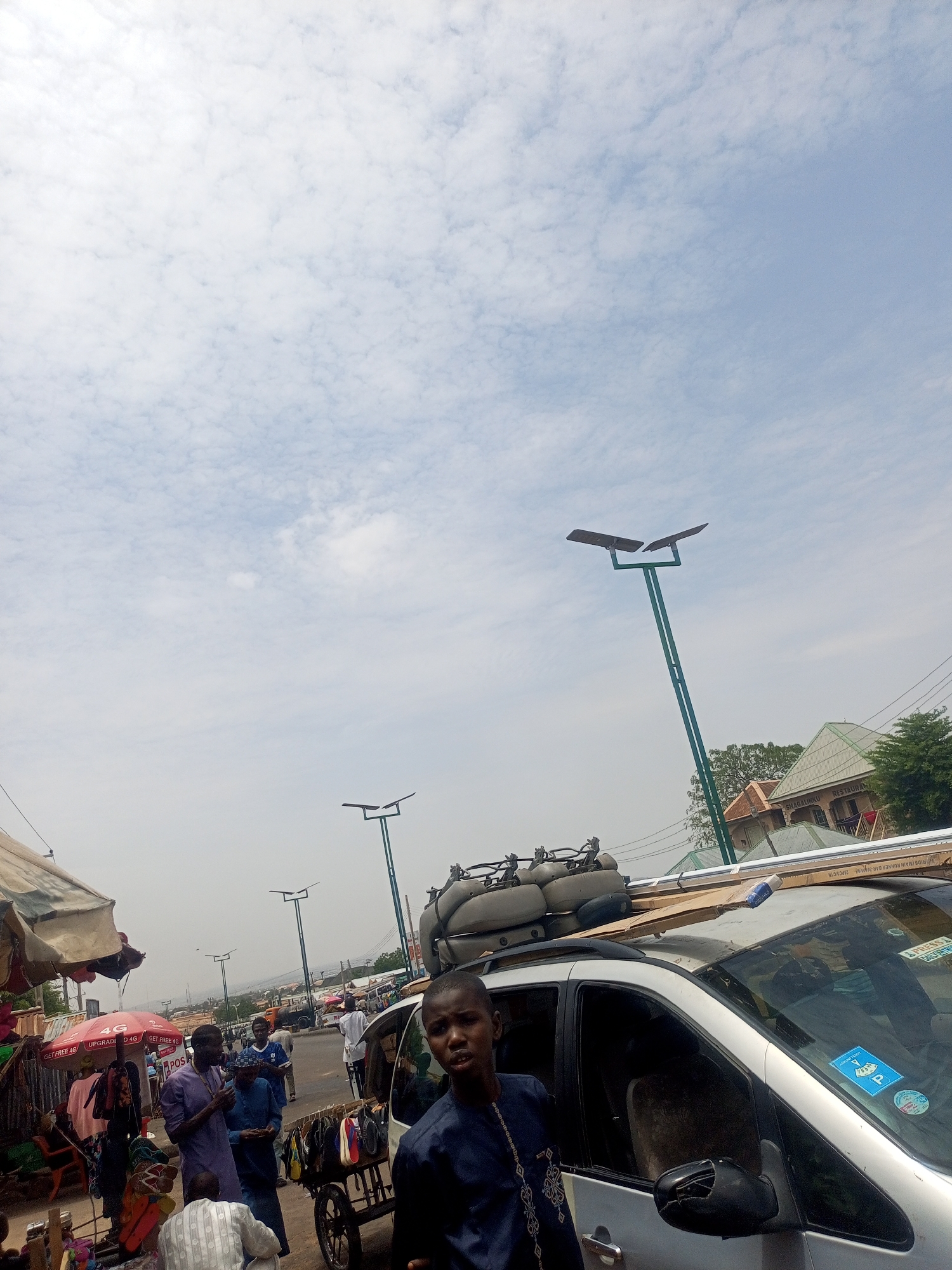 The image, taken during the day in an outdoor setting, shows a bustling scene that appears to be a marketplace in the city of Gombe, Nigeria. The sky is overcast with scattered clouds. In the foreground, a young man in a dark blue patterned tunic stands near the passenger side of a silver minivan. The van's roof is loaded with a variety of objects including inflatable floats, building materials, and other items secured by a rack.
To the left of the van, there's an active marketplace area with people and vendors displaying goods under a red umbrella and other makeshift canopies. Several individuals are dressed in traditional clothing, including purple and white garments. Some people are engaged in buying, selling, or conversation.
In the background, a row of tall, green street lamps with horizontally mounted lights stretches into the distance along the road. Buildings with green-painted roofs are visible among the trees. There is a restaurant sign in the background as well.