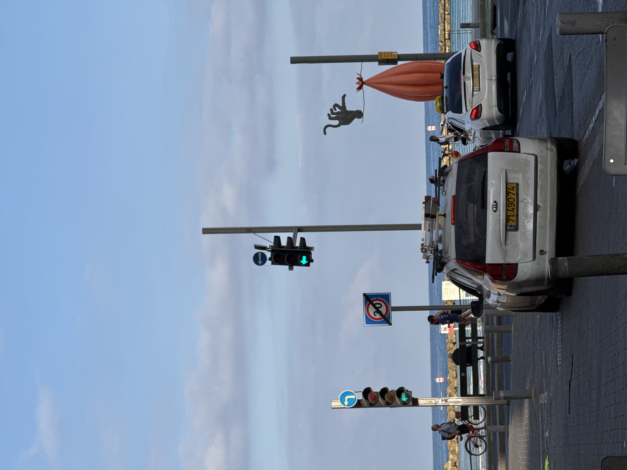 The image depicts a street scene in Tel Aviv, Israel, near the coastline. The foreground features a paved sidewalk and a street with two white cars. The car on the left is a white Kia station wagon with a yellow license plate reading "71-490-74". The car on the right is a white Mercedes sedan, likely a taxi, with a yellow taxi sign on its roof and a license plate reading "61-006-26". Traffic lights are visible; one on the left is green with a left-turn arrow, and another further back is also green. A speed limit sign with a red circle and a "30" inside crossed out by a red bar is also present, indicating no entry for vehicles exceeding 30.
In the middle ground, several people are visible. A man on a bicycle with a red frame stands near a bench by the sea. A man sits on the bench, facing away from the camera. Two individuals are jogging along the promenade. Behind them, a rocky breakwater separates the street from the sea.
The background is dominated by the blue sea and a partly cloudy sky. On the right side of the image, a distinctive orange, bulbous sculpture is mounted on a pole, with a dark silhouette of a figure with multiple limbs perched above it. The overall atmosphere suggests a late afternoon or early evening setting, with the sun casting a soft light.