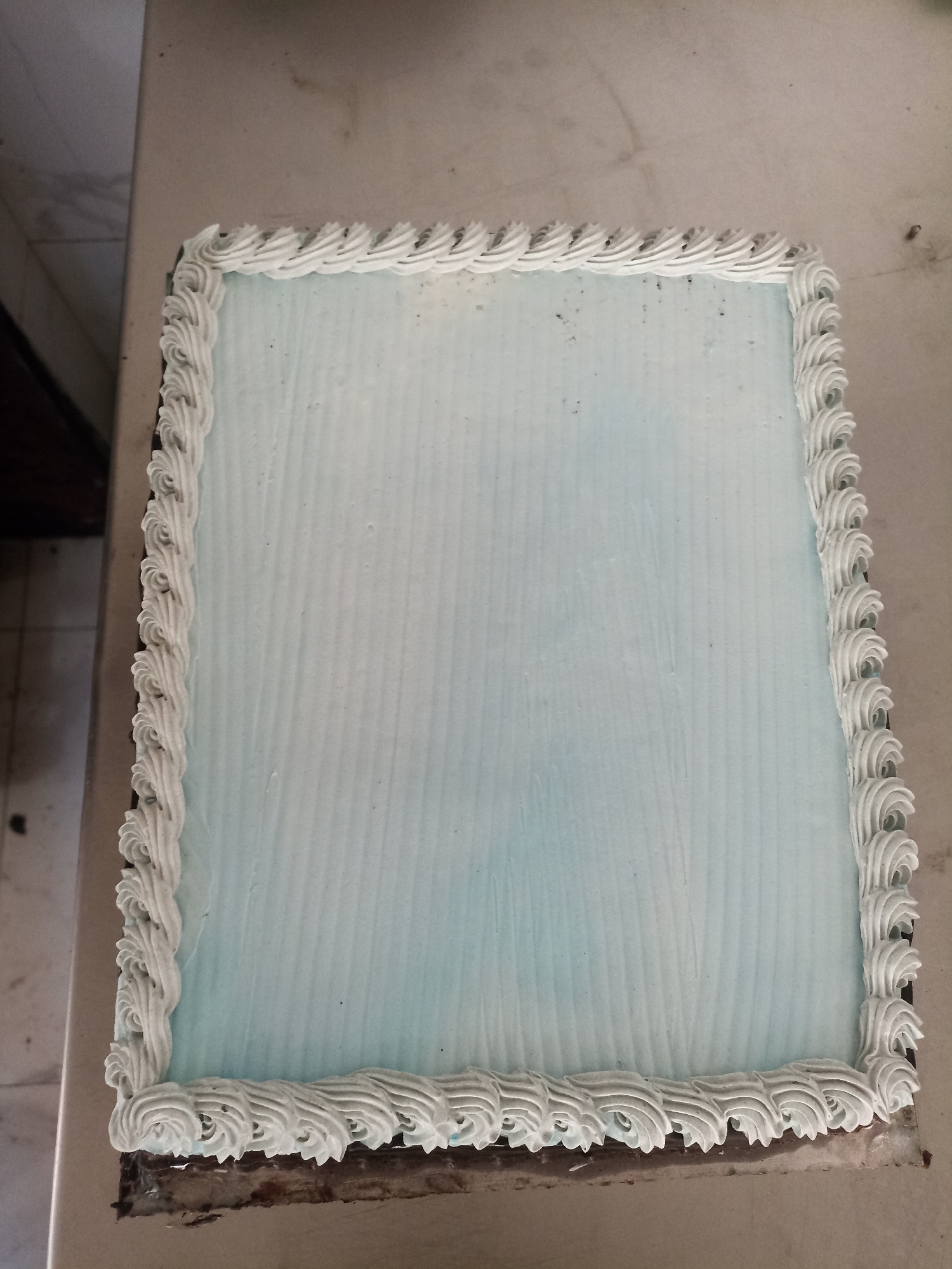 A rectangular cake with light blue frosting and white piped borders sits on a beige surface. The frosting has a ridged texture. The cake appears to be on a serving tray or board, with some of the underlying cake visible at the bottom edge, showing dark brown layers. The background is out of focus, with hints of a tiled wall and furniture on the left side. The overall scene is well-lit, suggesting an indoor setting. The image quality is decent, with clear focus on the cake. There are no people or animals visible in the image.
