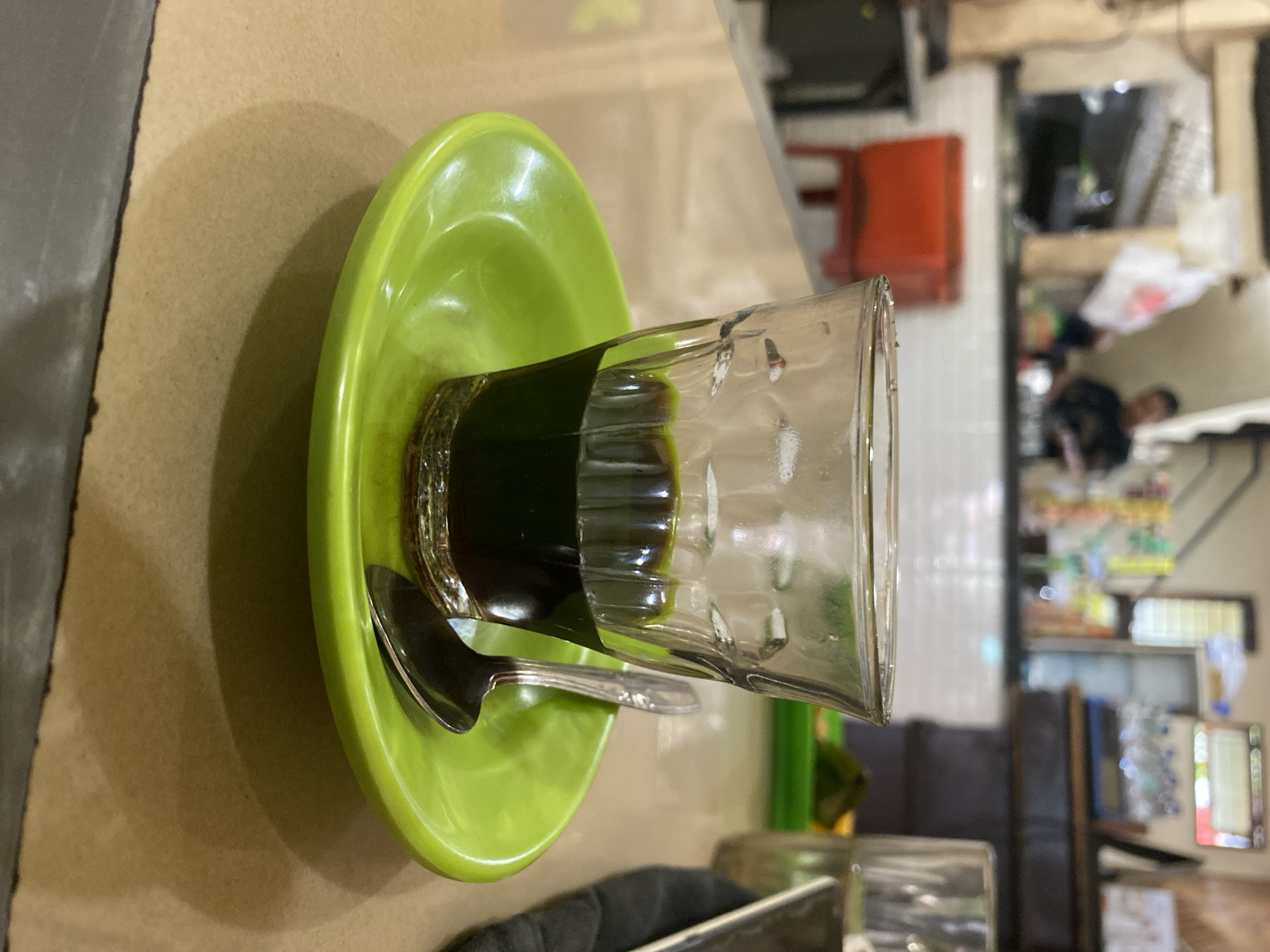 A close-up, eye-level shot shows a small glass of dark liquid, possibly coffee or tea, on a bright green saucer. A silver spoon rests on the saucer, slightly angled. The glass and saucer are placed on a light brown, textured surface, likely a table. In the blurred background, a casual eatery setting is visible, with a person standing behind a counter, and various items like a red cooler, bottles, and what appears to be a menu or display board. The overall atmosphere is that of a local establishment in Lhokseumawe, Indonesia.