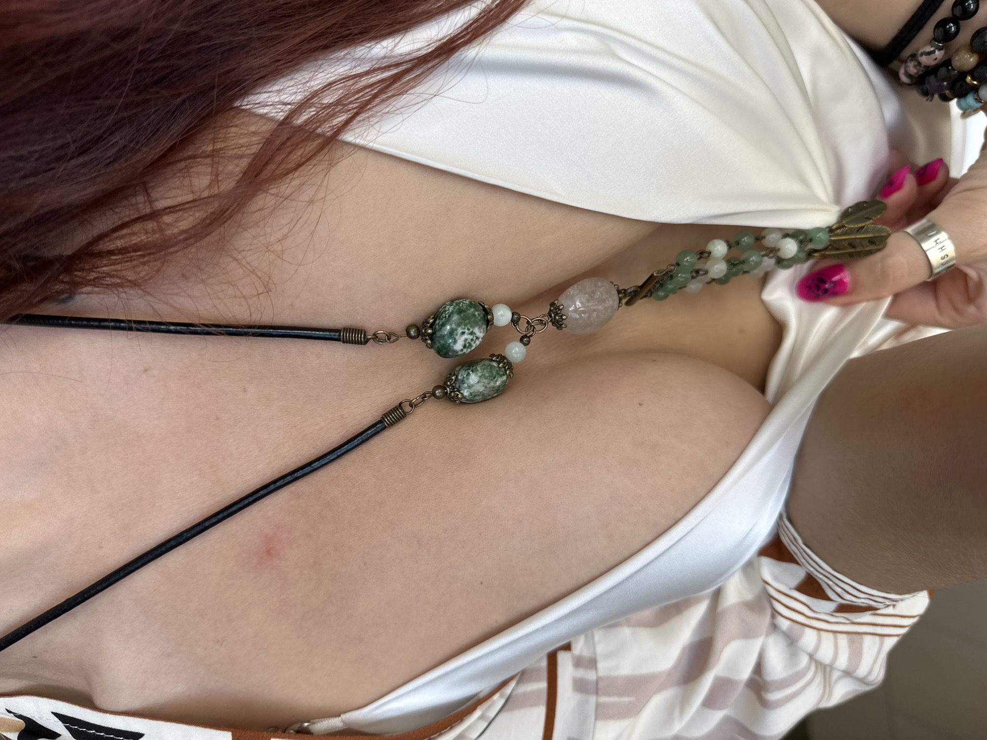 A close-up, high-angle shot captures the upper body of a person, likely female, within what appears to be an indoor setting in Vladimir, Russia. The person has long, reddish-brown hair cascading over their shoulder and back. They are wearing a white, satiny garment, possibly a top or dress, revealing a décolletage. Beneath or as part of the outfit, a second garment with brown and white stripes is visible.
A prominent feature is a decorative necklace with green, white, and translucent pink beads, strung on a black cord or leather strap. The necklace includes bronze-toned metal accents, some shaped like small feathers. The person's right hand is visible, adorned with bright pink nail polish, some featuring intricate designs. This hand is positioned near the necklace and garment, possibly adjusting or showcasing them. On the ring finger of this hand, a silver-colored ring is worn, engraved with the visible text "HHSA". Additional bracelets made of black and multi-colored beads are visible on the wrist. The lighting is soft and even, suggesting an interior environment without direct sunlight. The image focuses on textures, colors, and personal accessories.