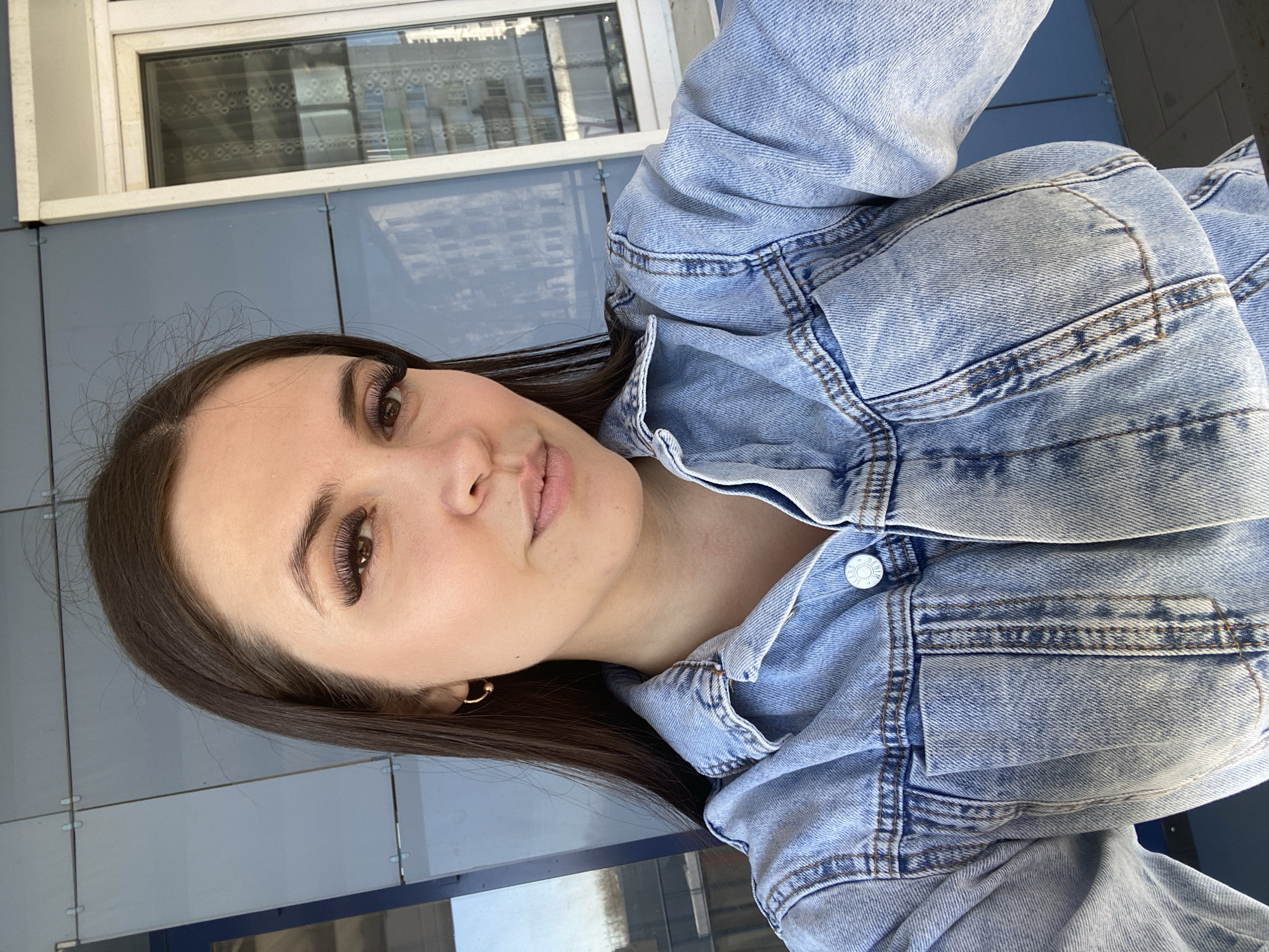A selfie of a woman with dark hair and brown eyes wearing a light blue denim jacket. She has false eyelashes and is wearing small hoop earrings. The background shows a light blue building exterior with a window reflecting the city buildings. The location is likely in or near Chaikovsky, Russia.