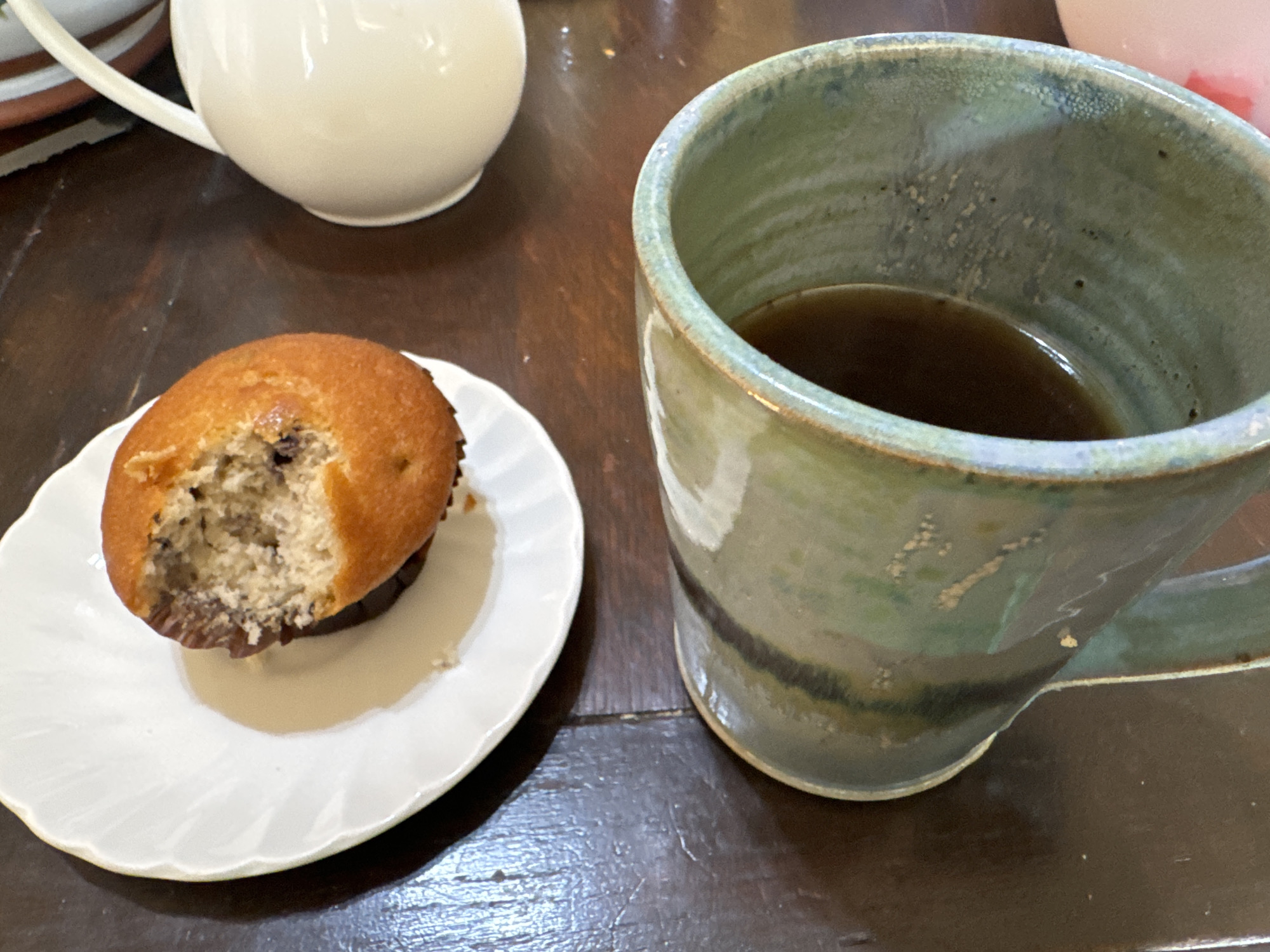 The image captures a close-up, overhead view of a dark wooden table. On the table, a white scalloped plate holds a partially eaten blueberry muffin. To the right of the plate, a white teapot with a curved handle is visible. In the foreground, a large, green ceramic mug filled with dark liquid, likely coffee or tea, dominates the lower portion of the frame. The mug has a textured, earthy glaze with variations in green and darker tones. The scene appears to be set indoors, possibly in a cafe or home setting, and there is no indication of the city of Waterford, Ireland.