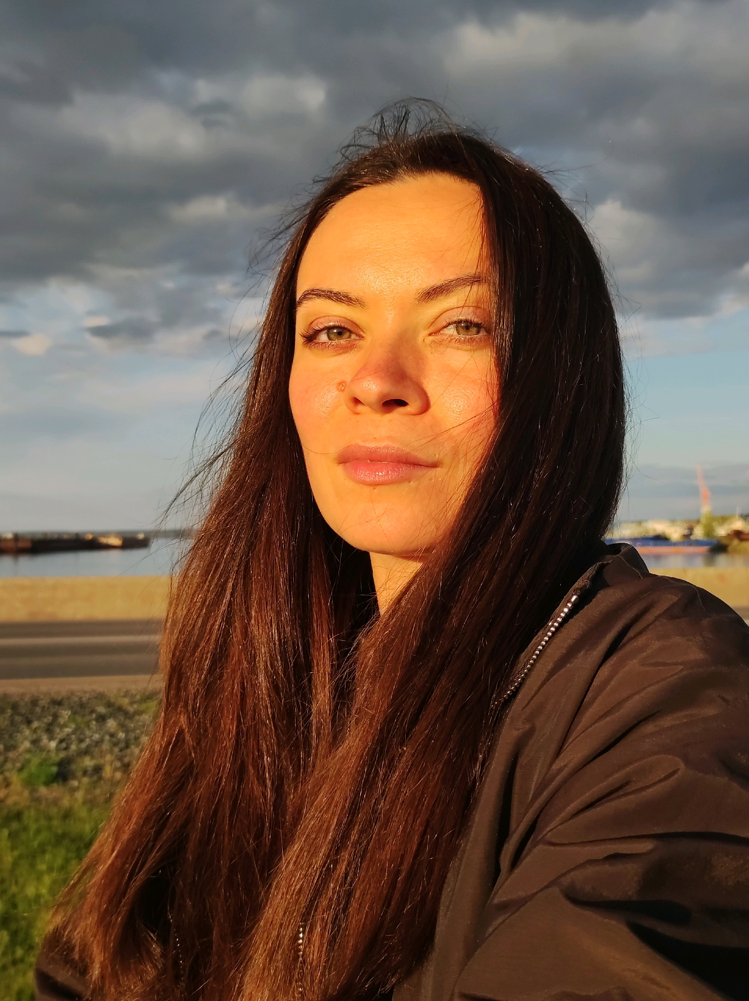 A woman is photographed in a close-up, selfie-style shot, facing the camera. She has long, dark brown hair that is slightly windswept and falls over her shoulders and onto her black jacket. Her eyes are open and looking directly at the viewer, and she has light skin with a reddish hue, possibly from the setting sun. The background suggests an outdoor location with a body of water, a road, and cloudy skies. Specifically, the image appears to be taken in Chaikovsky, Russia, given the presence of the Kama River which forms the eastern boundary of the city, and the industrial infrastructure visible across the water, indicative of the city's port and power plant. The lighting suggests it is either sunrise or sunset, casting a warm glow on the woman's face.