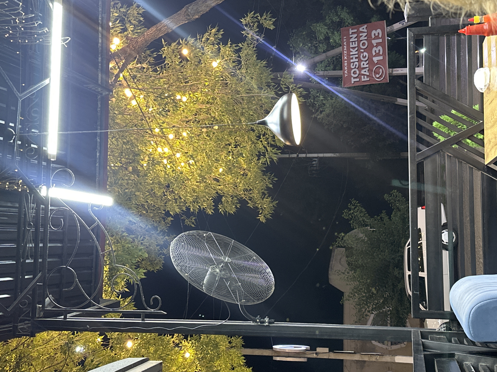 The image depicts an outdoor seating area at night, likely a cafe or restaurant, with a clear view of the street and surrounding environment in Fergana City, Uzbekistan. A large industrial fan is mounted on a pole to the left, and a decorative pendant light hangs from above the seating area. The area is illuminated by string lights woven through the branches of a tree and a long fluorescent light fixture mounted overhead. A white car is parked on the street in the background, and a sign for "TAKSI XIZMATI TOSHKENT FARGO'ONA 1313" is visible on a pole. The foreground shows a portion of the outdoor seating furniture, including a blue chair and a wooden table with a few items on it, possibly condiments. The overall scene is one of a public space at night, illuminated and active with the presence of vehicles and signage indicating local services.