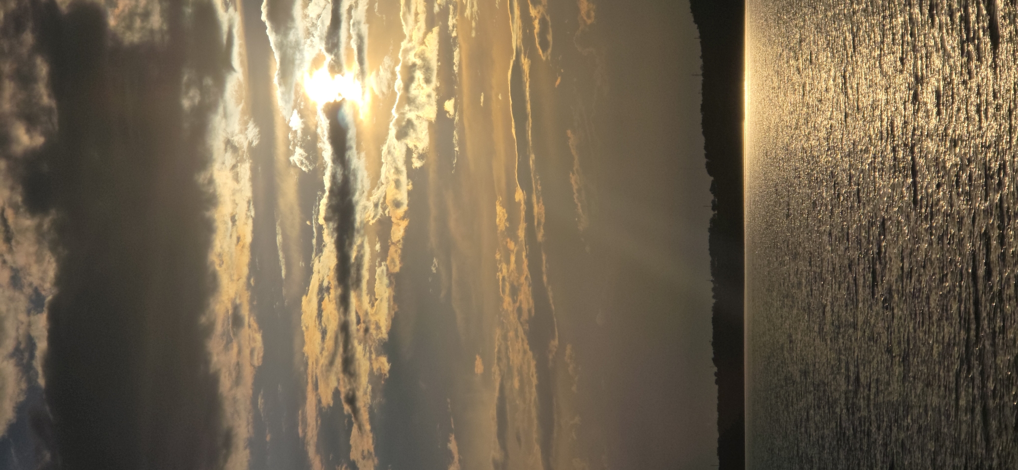 The image depicts a serene lake at sunset, with a dark, silhouetted treeline forming the horizon. The sky is filled with dramatic, textured clouds, illuminated by the golden rays of the setting sun which create a shimmering path of light across the water. The scene is devoid of any human presence or activity. The location is identified as Pristan'-Przheval'sk, Kyrgyzstan.