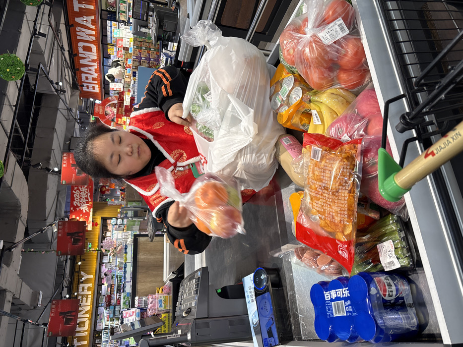 The image depicts a transaction at a supermarket checkout lane in Jiangjin, China. A person, appearing to be female with dark hair and wearing a black long-sleeved shirt under a red, embroidered vest with a white collar, is standing behind a metallic gray counter. She is actively transferring items, holding a clear plastic bag containing red and green fruit in her left hand, while her right hand is inside a larger white plastic bag containing other groceries. Her gaze is directed downwards at the items.
On the checkout counter, a variety of groceries are visible, including blue multipacks of canned Pepsi beverages, multiple clear plastic-wrapped packages of processed meats, two clear plastic bags filled with red tomatoes with attached price labels, a bunch of yellow bananas, and green vegetables in a plastic bag. To the left, a black cash register terminal with a keyboard and screen is present, alongside a blue device possibly for mobile payments or scanning, displaying partially visible Chinese text "解锁手机" (unlock phone). The handle of a black shopping cart, bearing the "JIAJIA XIN" logo and text, extends from the bottom right foreground.
The background reveals a brightly lit supermarket interior with rows of shelves and various product displays. Overhead signage includes "STATIONERY" on the left and partially visible "BRAND WATER" on the right. Red banners with Chinese text, notably "2026 新年快乐" (2026 Happy New Year), hang from the ceiling, which also features exposed ducts, lighting fixtures, and decorative green spherical objects.