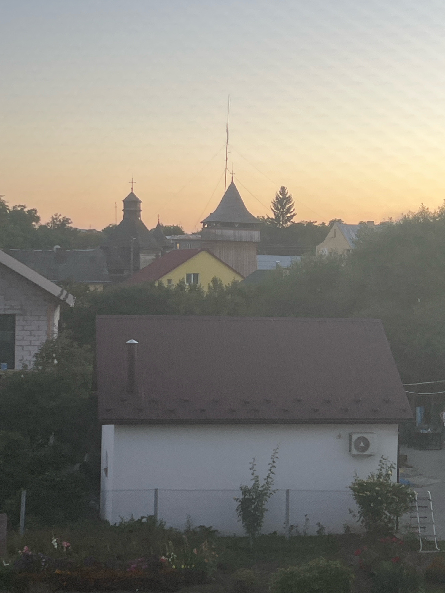 The image shows a residential area at dusk in Drohobych, Ukraine. In the foreground, a white house with a dark roof is visible, featuring an air conditioning unit on its exterior wall. A small garden with flowers and bushes is situated in front of the house. Beyond this, a cluster of buildings and trees creates a layered landscape. In the middle ground, a yellow house with a red roof stands out, surrounded by greenery. Further in the background, the distinct spires of what appear to be churches or religious buildings are silhouetted against the pale, warm-toned sky, characteristic of a sunset or sunrise. A tall, slender antenna or spire rises above the rooftops. The overall scene depicts a quiet, possibly rural or suburban, setting in Ukraine during the evening or early morning light.