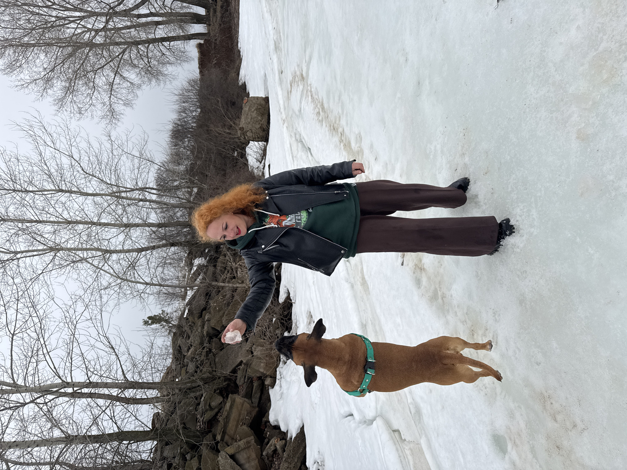 A red-haired person in a black leather jacket, green sweatshirt, and brown pants stands on a snow-covered area with a brown dog, possibly a Belgian Malinois, in a green harness. The person holds a piece of ice or snow in their outstretched hand, seemingly offering it to the dog. The environment consists of a rocky outcropping and bare trees. The image appears to have been taken in the city of Chaikovsky, Russia.