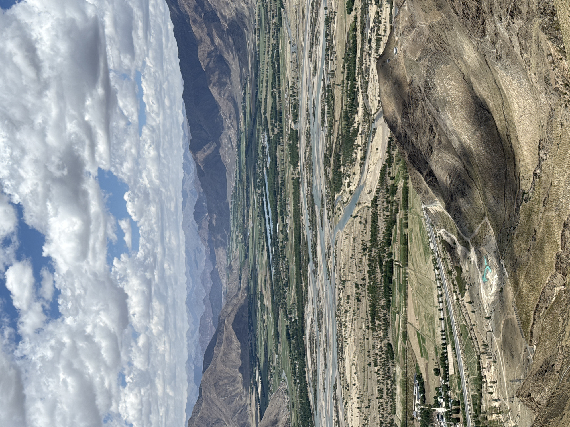 The image depicts a valley in Lhasa, China, viewed from an elevated perspective. The foreground features a dry, mountainous terrain transitioning into a valley with a braided river system. The river separates green agricultural fields and sparsely vegetated areas. A road runs parallel to the river through the valley. The background shows layered mountains receding into the distance, with a sky filled with cumulus clouds overhead.