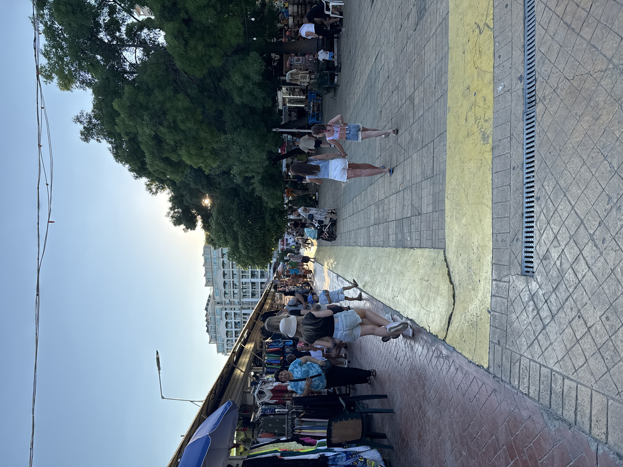 The image depicts an outdoor scene on a pedestrian walkway, likely in Nesebar, Bulgaria. The perspective is tilted, making it appear as though the viewer is looking down at the scene. In the foreground, a market stall displays various clothing items laid out on tables and hung on racks. A woman wearing a blue patterned shirt sits behind one of the tables, appearing to be a vendor.
Further down the walkway, several people are walking and browsing. Two young women in casual summer attire, shorts and tops, are walking away from the camera, while another woman in denim shorts and a patterned top walks towards them. Behind them, more people are visible, some seated at tables under trees, suggesting a cafe or restaurant area. The sunlight filtering through the trees indicates it is daytime, likely late afternoon or early evening given the warm light. The sky is clear and blue.
The walkway is paved with interlocking stones, with a distinct yellow stripe running along the edge of the market area. A drainage grate is visible along the right side of the walkway. In the background, a multi-story building with balconies and white facades suggests an urban or resort setting. The overall atmosphere seems relaxed and leisurely, typical of a tourist area. No visible text is discernible in the image.
