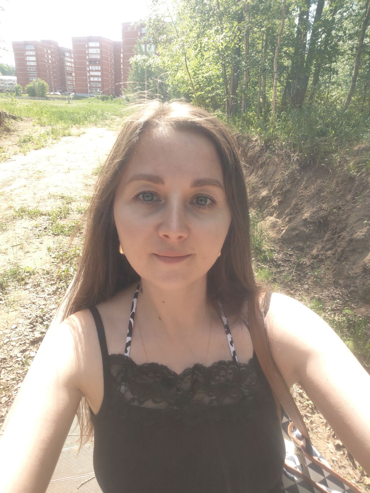 A woman is standing outdoors on what appears to be a dirt path or clearing. Behind her to the left, several multi-story brick apartment buildings are visible in the distance, suggesting an urban or suburban location. A wooded area with trees and foliage is present to the right of the woman. The woman is wearing a black top with lace trim, and her long brown hair frames her face. She is facing the camera, giving the impression of a self-portrait or selfie. The sunlight is bright, indicating a sunny day.