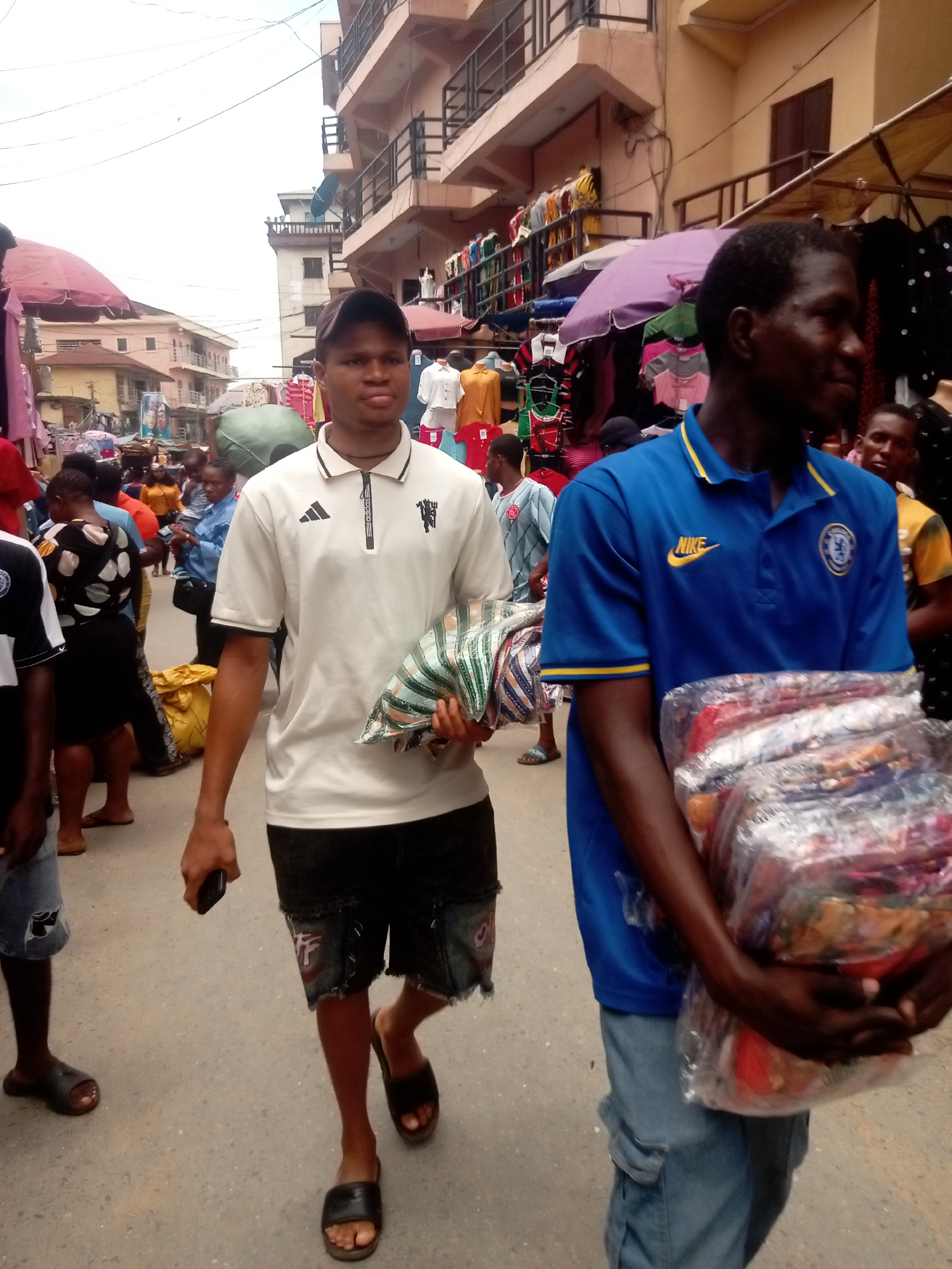 The image shows a bustling street scene in Fegge, Nigeria. Two men are the primary subjects: The man on the left is wearing a white polo shirt with black trim, dark denim shorts, a black baseball cap, and sandals, carrying a striped bag. The man on the right is wearing a blue Nike/Chelsea soccer jersey and light-wash denim cargo pants, carrying a large package wrapped in clear plastic. The background contains a crowd of people, vendor stalls with clothing, and multi-story buildings. The image has overcast lighting.
