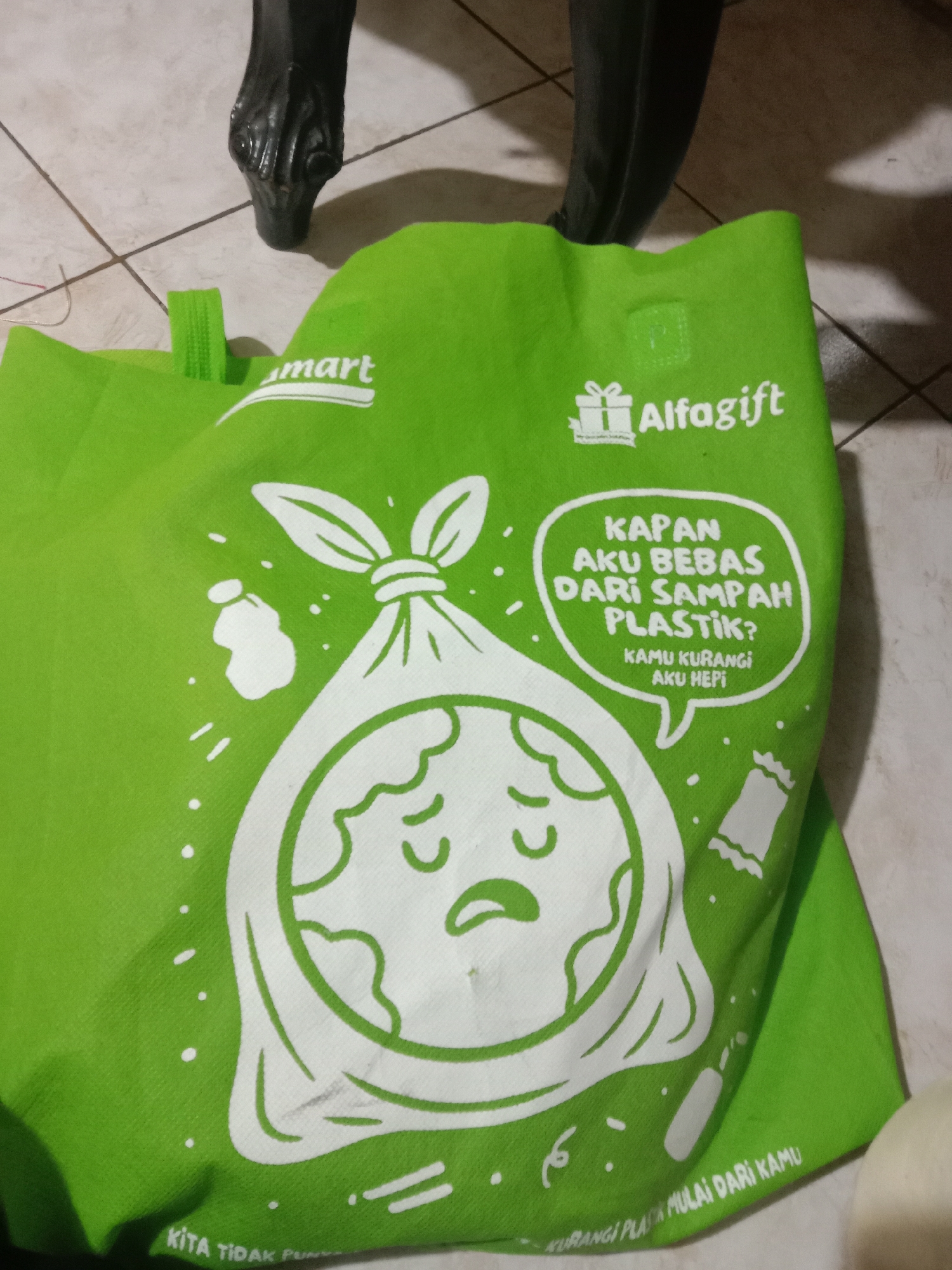 The image shows a bright green reusable shopping bag with a white graphic design and text. The bag is lying on a tiled floor, with the leg of a dark-colored piece of furniture visible in the upper portion of the image. The graphic on the bag depicts a sad-looking Earth inside a trash bag, with text in Indonesian. The text translates to "When will I be free from plastic waste? You reduce, I'm happy." and "Let's start reducing plastic from you." The bag appears to be a promotional item from "Alfamart" and "Alfalifegift." The location is not specified in the image, so it cannot be determined if it is in Madiun, Indonesia.