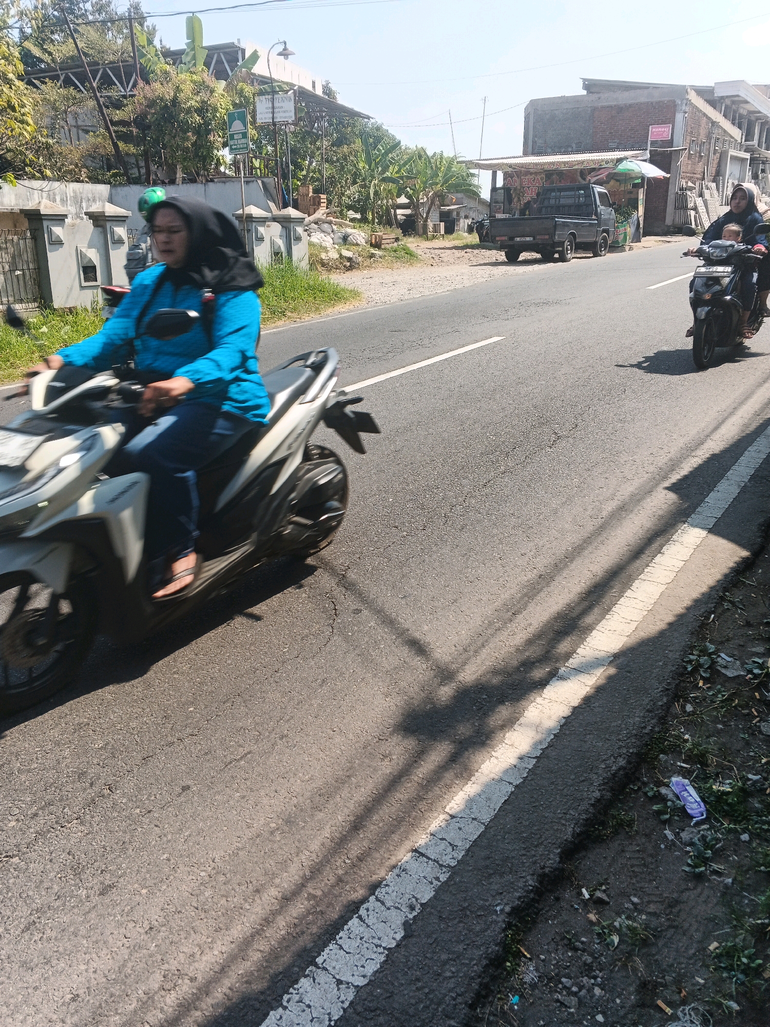 A medium shot captured on a sunny day shows a paved road with a white stripe marking the lane. On the left, a woman wearing a blue long-sleeved shirt and a black hijab rides a white scooter. She is leaning forward, her hands on the handlebars, and her face is blurred due to motion. Her legs are visible, wearing dark pants and sandals. A shadow of the scooter and its rider is cast on the road. Further down the road, another scooter with two riders, a woman and a child, travels in the same direction. They are partially visible. In the background, there are buildings, trees, and a parked black pickup truck. The scene is set in Bandungan, Indonesia, indicated by the context provided. The environment appears to be a rural or semi-urban area.