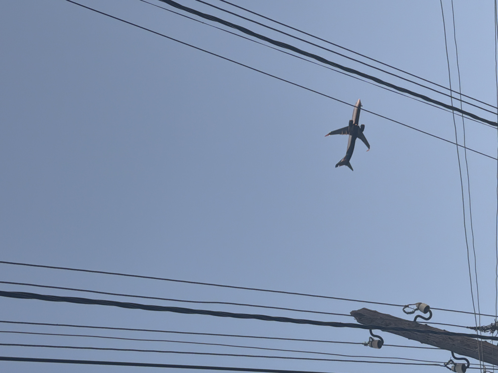 The image depicts a clear blue sky with a commercial jet airplane flying overhead. Electrical wires are visible in the foreground, running horizontally across the frame, and a wooden utility pole with insulators is in the lower right. No people are visible. The location, Yerevan, Armenia, is not determinable from the image.