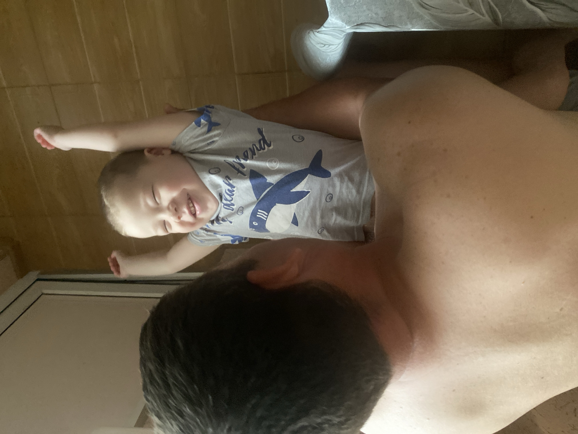 The image shows a fair-skinned adult with short dark hair, and a young child with blond hair. The child is wearing a grey t-shirt with a shark design and appears to be smiling. The adult is shirtless. The background appears to be a bathroom with tiled walls and a shower door visible. The location is not discernible from the image.