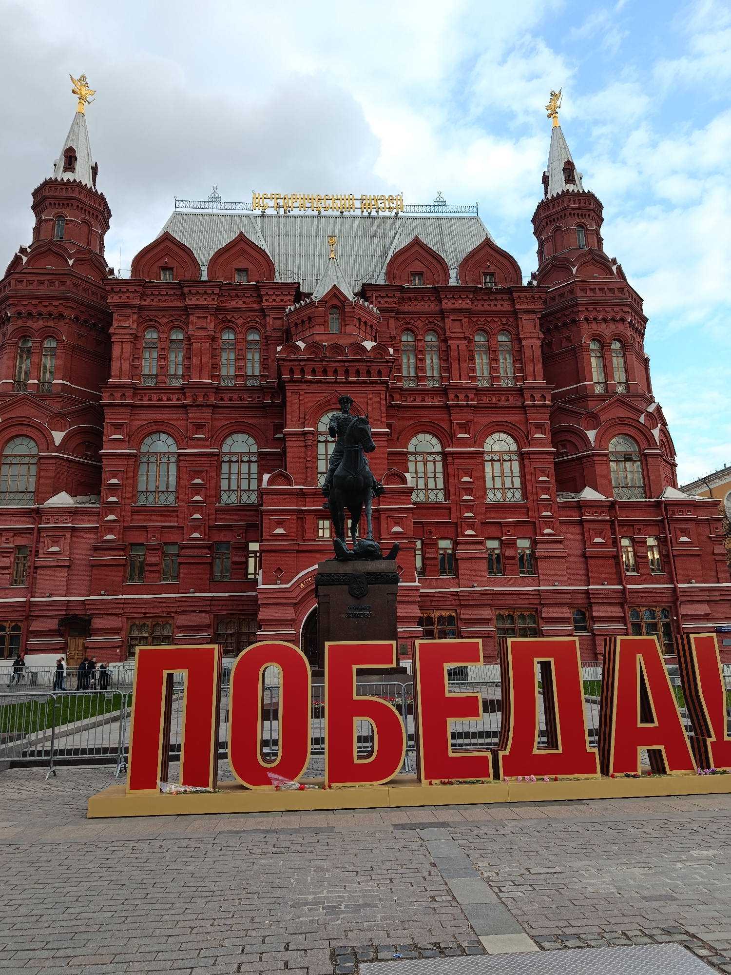 The image shows the State Historical Museum in Moscow, Russia, with a large red sign in front that says "ПОБЕДА!" (Victory!). The museum is a large red brick building with white trim and multiple towers and windows. A statue of a rider on a horse is in front of the museum entrance. A metal fence is in the background near the front of the museum, and a few people are standing nearby. The ground is paved with gray bricks. The sky is cloudy with patches of blue.