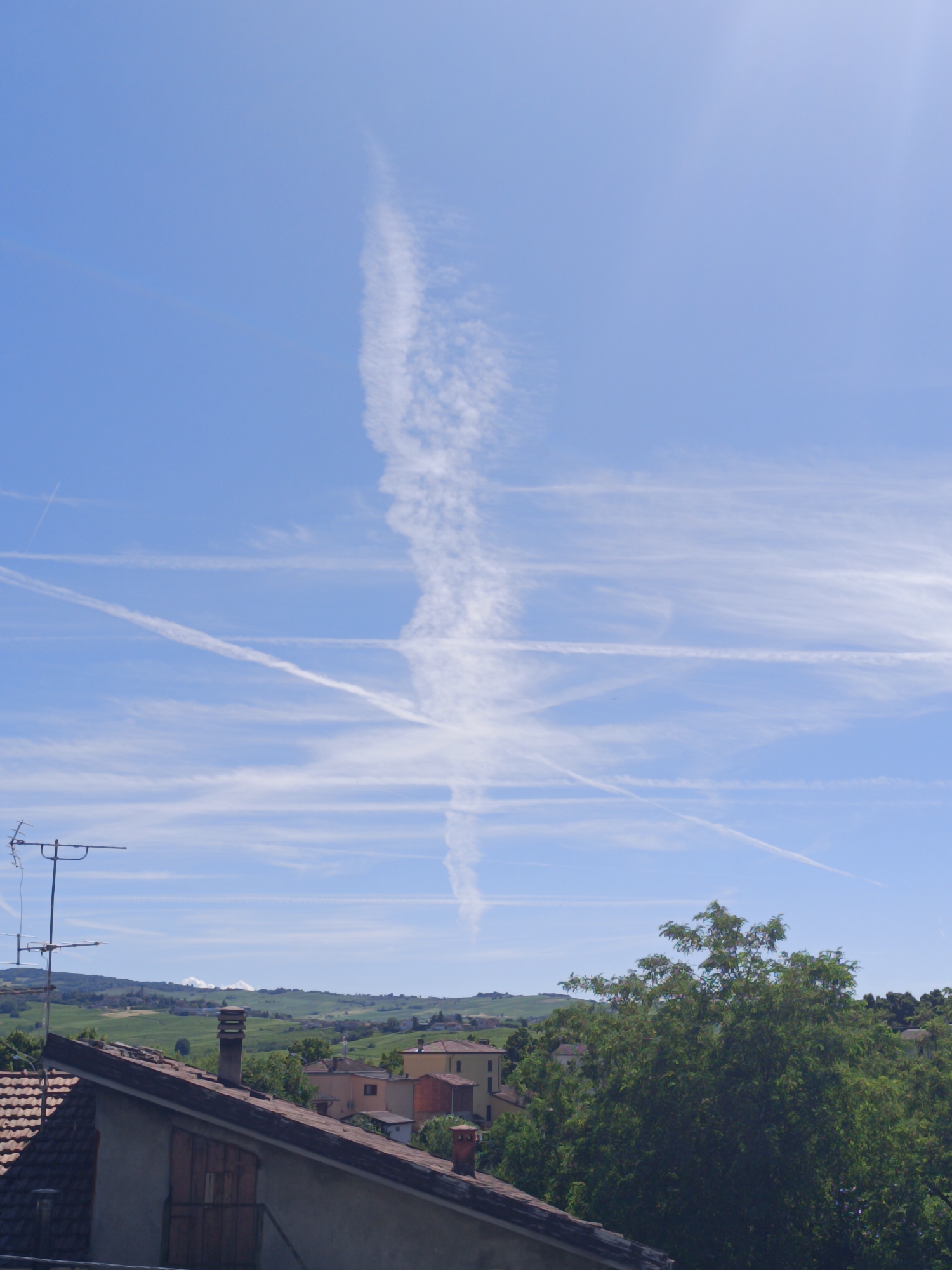 A daytime outdoor scene captures a panoramic view from an elevated position, looking over rooftops towards a distant, hilly landscape under a vast blue sky. The sky, which occupies the upper two-thirds of the image, is a prominent feature, displaying numerous white contrails from aircraft. One particularly long, thick, and somewhat diffused contrail stretches vertically down the center-right, while several other thinner, linear contrails crisscross the sky horizontally and diagonally.
In the foreground, the sloping terracotta-tiled roofs of residential buildings are visible. A silver-colored rooftop antenna with multiple elements is prominently situated on the left, extending above a roof. Two chimneys, one slender and dark, the other shorter and reddish-brown, rise from different rooftops.
The midground reveals more houses with light-colored walls and reddish roofs, nestled amongst dense green trees. A large, leafy green tree dominates the lower right corner, partially obscuring elements behind it.
The background transitions into a series of gently rolling hills, covered in lush green vegetation, possibly agricultural fields or vineyards. Clusters of buildings or small villages can be seen on the distant hillsides. The clear blue sky and bright lighting indicate it is daytime, likely morning or early afternoon. No people or visible text are present in the image.