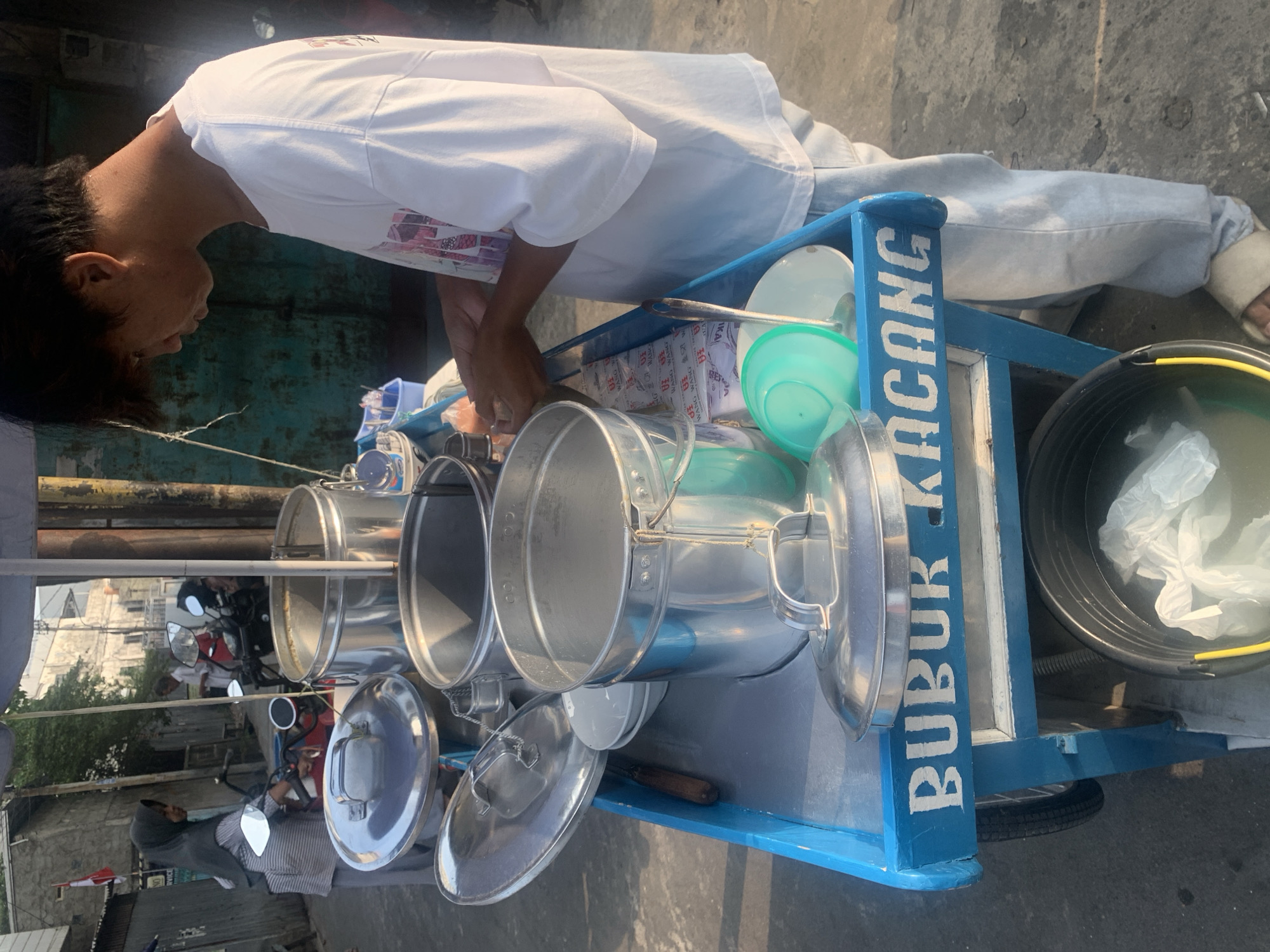 A young man in a white t-shirt and light blue pants stands behind a blue food cart. The cart has the words "BUBUR KACANG" written on its side. Several metal pots, bowls, and a ladle are visible on the cart. In the background, a woman wearing a grey hijab and striped shirt is standing near a motorcycle, and other buildings and a red flag are visible. The scene appears to be in a street in Palembang, Indonesia.