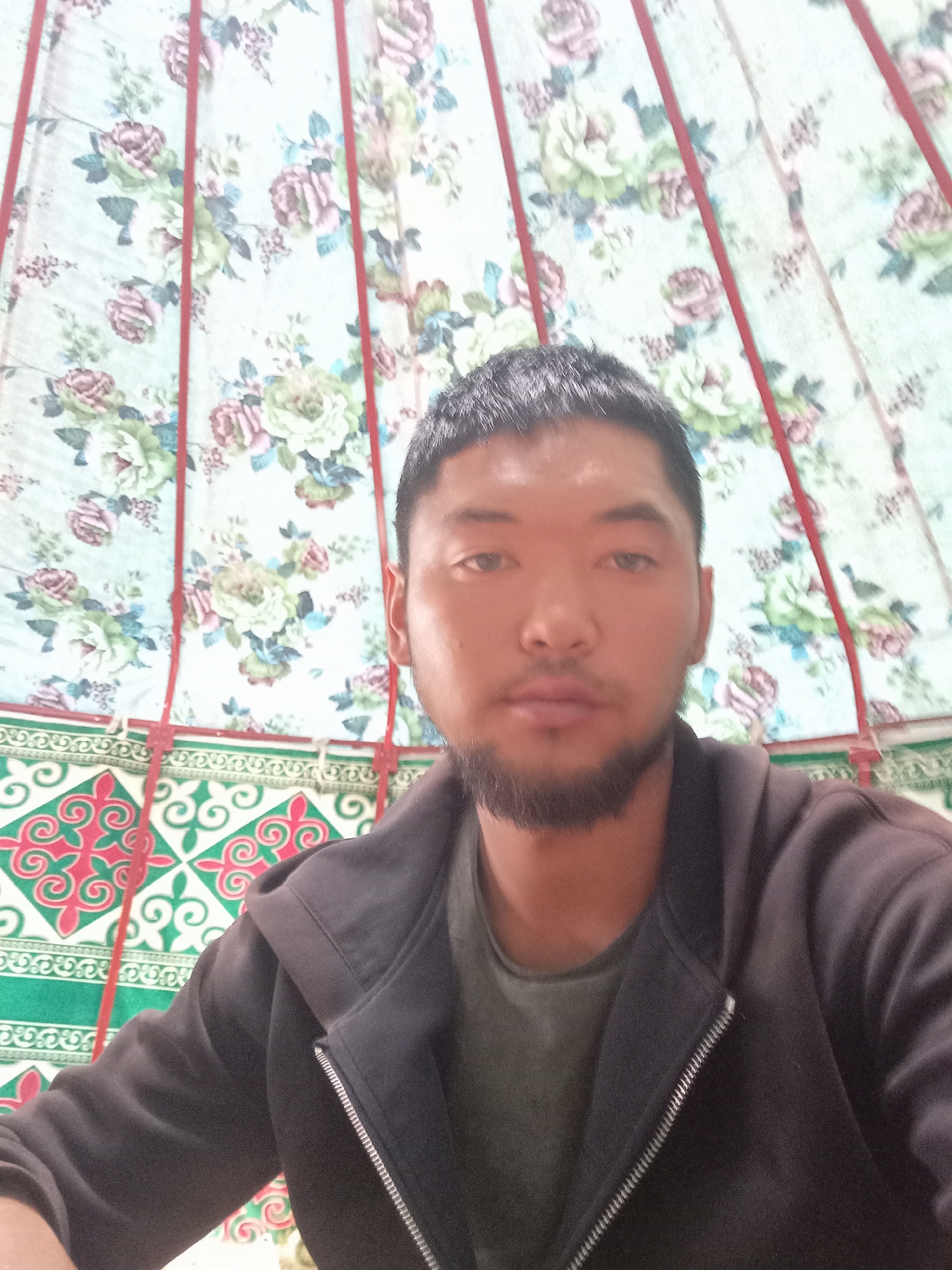 A close-up, medium shot captures a young man with short dark hair, a neatly trimmed beard, and dark eyes looking directly at the camera. He is wearing a dark hooded jacket over a dark green t-shirt. The background consists of the interior of a yurt, with floral patterned fabric walls adorned with red vertical supports. Below the floral fabric, there is a green fabric border with a geometric pattern of red and white swirling designs. The lighting appears to be natural and even, suggesting an indoor setting within a yurt in Bayet, Kyrgyzstan.