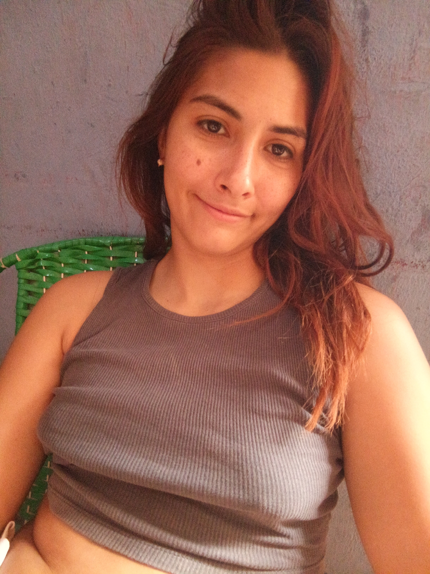 A medium shot captures a fair-skinned woman with reddish-brown hair and brown eyes, who is seated. She is wearing a gray tank top. Her hair is somewhat disheveled and falls around her face and shoulders. A portion of a green woven chair is visible behind her left shoulder. The background is a muted gray wall. The location in the city is Unknown, Unknown.
