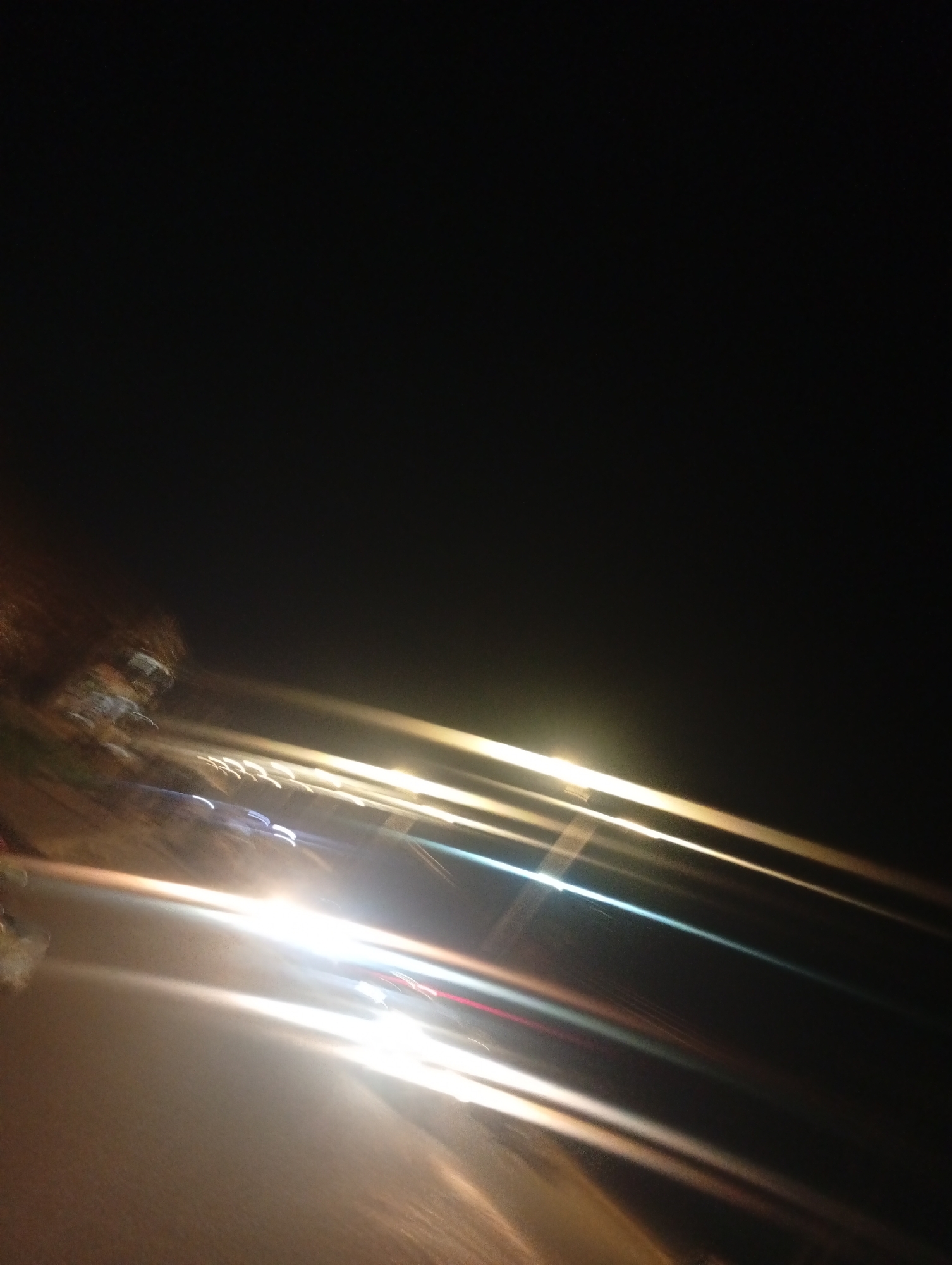 The image depicts a blurred nighttime scene, likely taken from a moving vehicle in Bukuru, Nigeria. Streaks of light suggest passing headlights and taillights on a road. The dark sky occupies the upper portion of the frame, while the lower half is dominated by the blurry road and artificial lights. There are no clearly discernible people or specific landmarks, emphasizing the impression of motion and low visibility. The overall impression is that of a fleeting glimpse of a nocturnal urban environment.