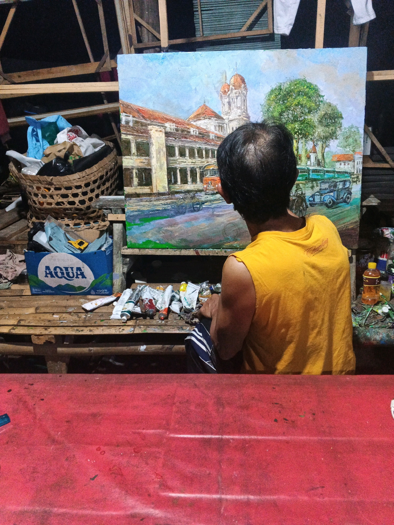 The image depicts an artist in Ungaran, Indonesia, working on a painting. The artist, seen from behind, is wearing a yellow sleeveless shirt and striped pants. They are seated on a wooden bench in what appears to be a rustic, possibly outdoor or workshop setting, with unfinished wooden structures visible in the background.
The painting on the easel shows a scene with classical architecture, including a building with arches and domes, and trees. Several vintage-looking vehicles are also depicted. In front of the artist, on the wooden bench, are tubes of paint, brushes, and a blue box labeled "AQUA," which is a popular Indonesian bottled water brand. A woven basket filled with various items, including plastic bags, is also present on the bench.
The overall environment suggests a creative space, possibly an artist's studio or a temporary setup for painting. The lighting is dim, creating a moody atmosphere. The presence of the AQUA box and the surrounding wooden structures might indicate a location in a less urbanized area or a traditional workshop in Ungaran.