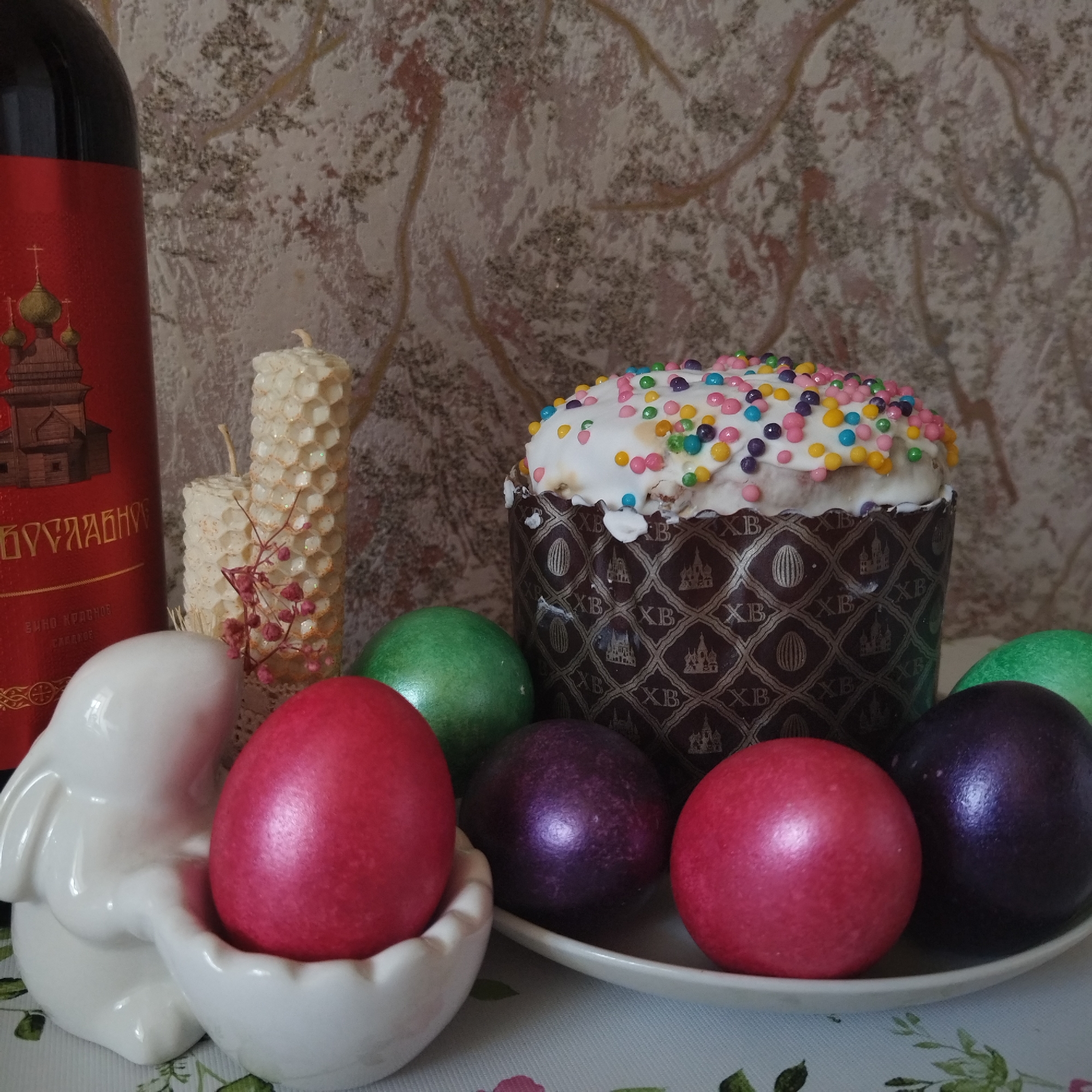 This media file captures a festive still life arrangement, strongly indicative of an Orthodox Easter celebration, set indoors on a table.
In the foreground, a white ceramic bunny figurine acts as an egg holder for a vibrant, shimmering red Easter egg. To its left, a dark red bottle, likely wine, features a label with a detailed illustration of a church adorned with golden onion domes. Visible text on the label includes "БОГУСЛАВНОВ" (Boguslavnov), "ВИНО КРАСНОЕ" (Red Wine), and "СЛАДКОЕ" (Sweet).
Behind the bunny and bottle are two beeswax candles, one tall and cylindrical with a prominent honeycomb texture, and a shorter, similar candle, both decorated with small sprigs of dried reddish plant material.
Dominating the right side of the composition is a traditional kulich (Easter bread or cake). It sits in a decorative brown paper wrapper emblazoned with repeating patterns of "XB" (Cyrillic abbreviation for "Christ is Risen"), stylized Easter eggs, and church motifs. The kulich is generously topped with white icing and a colorful array of sprinkles in hues of yellow, pink, blue, and purple.
Arranged around the kulich and on a white plate are several more dyed Easter eggs, exhibiting a distinct metallic or pearlescent finish, in rich shades of green, purple, and red. The setup rests on a light-colored surface, possibly a tablecloth with a subtle leaf pattern, against a textured wall featuring a mottled pattern of light brown and gray. The lighting is soft and even, suggesting a daytime setting, and the overall mood is celebratory and traditional.