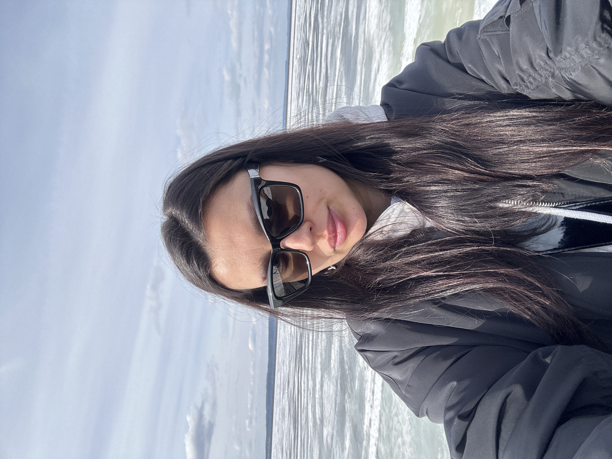 A woman with long dark hair and sunglasses is taking a selfie. She is wearing a black jacket over a white hooded sweatshirt. Behind her is a body of water, potentially a lake or river, with light-colored water under a partly cloudy sky. The location is likely Chaikovsky, Russia, given the context.