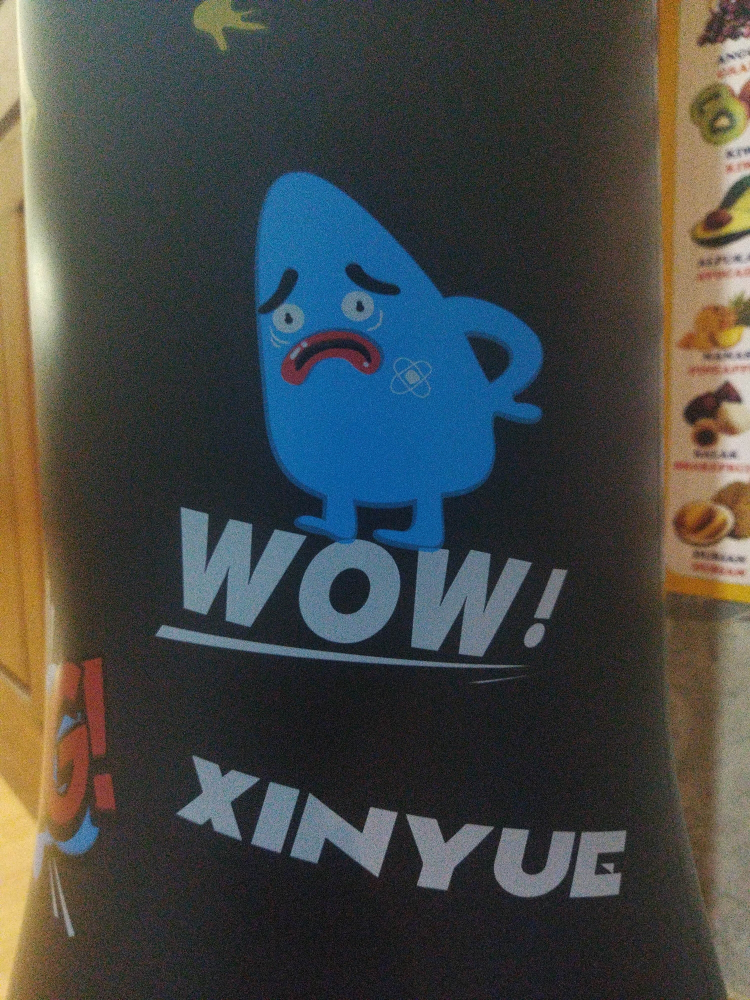 The image depicts a close-up of a dark-colored display stand, possibly for promotional purposes. Centered on the stand is a cartoonish blue character with a distressed facial expression, featuring large, round eyes and a downturned mouth. Below the character, the word "WOW!" is prominently displayed in large, white, block letters, underlined by a thin white line. Further down, the word "XINYUE" is written in similar white block letters. To the left of "XINYUE" is a red and blue graphic with stylized lines. On the right side of the image, a vertical strip displays images of various fruits and food items, each with a label in small text, which is largely illegible due to the image resolution. The lighting suggests an indoor environment, possibly a commercial space. There is no visible indication of its specific location in the city of Suci, Indonesia.