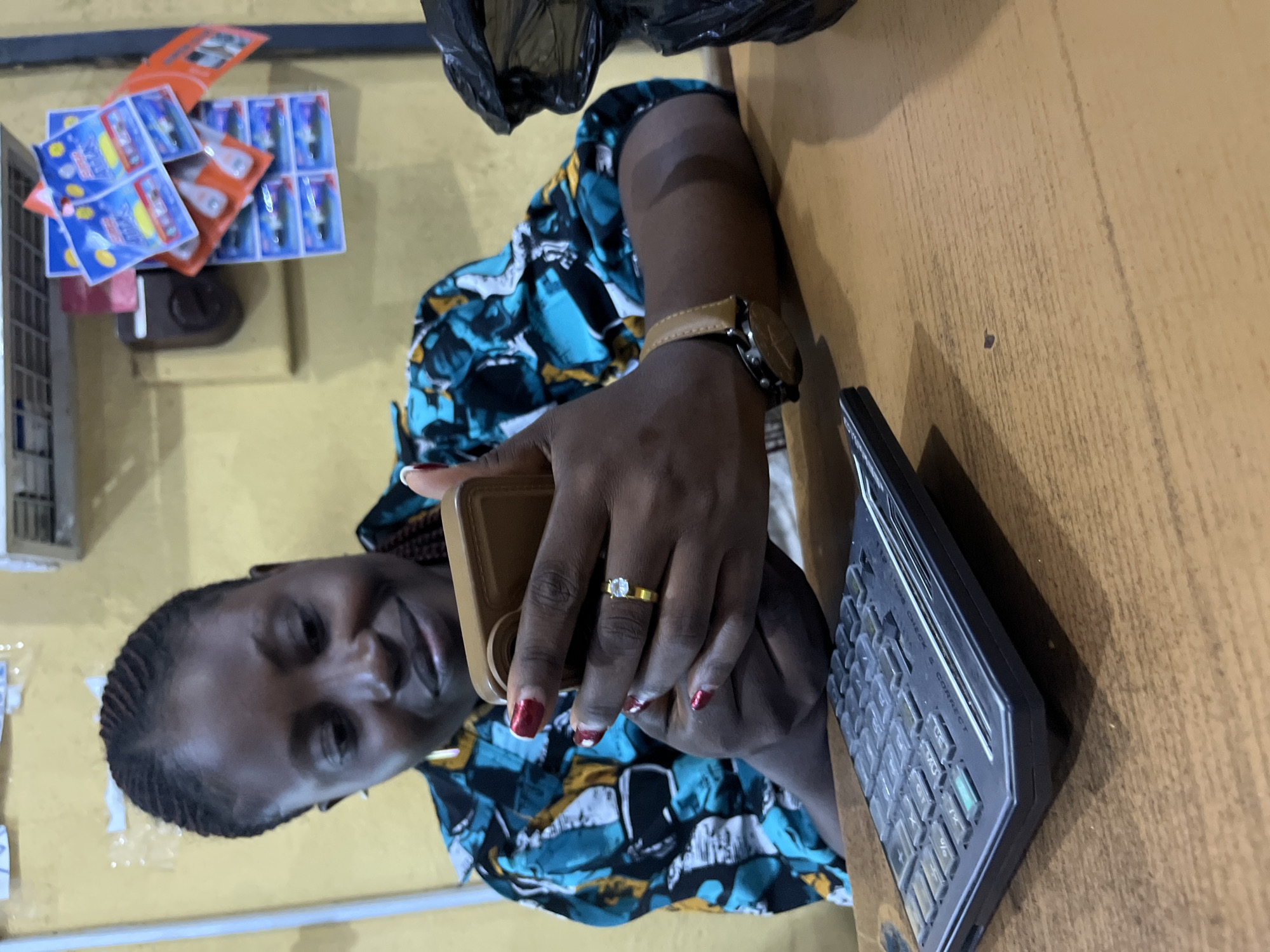 A Black woman with braided hair is looking down at a mobile phone in her hands. She is wearing a colorful shirt and a ring on her left hand. A calculator sits on the wooden table in front of her. The background is a yellow wall with some items hanging on it. The scene is indoors and appears to be in a shop or office. The location is the city of Bukuru, Nigeria.