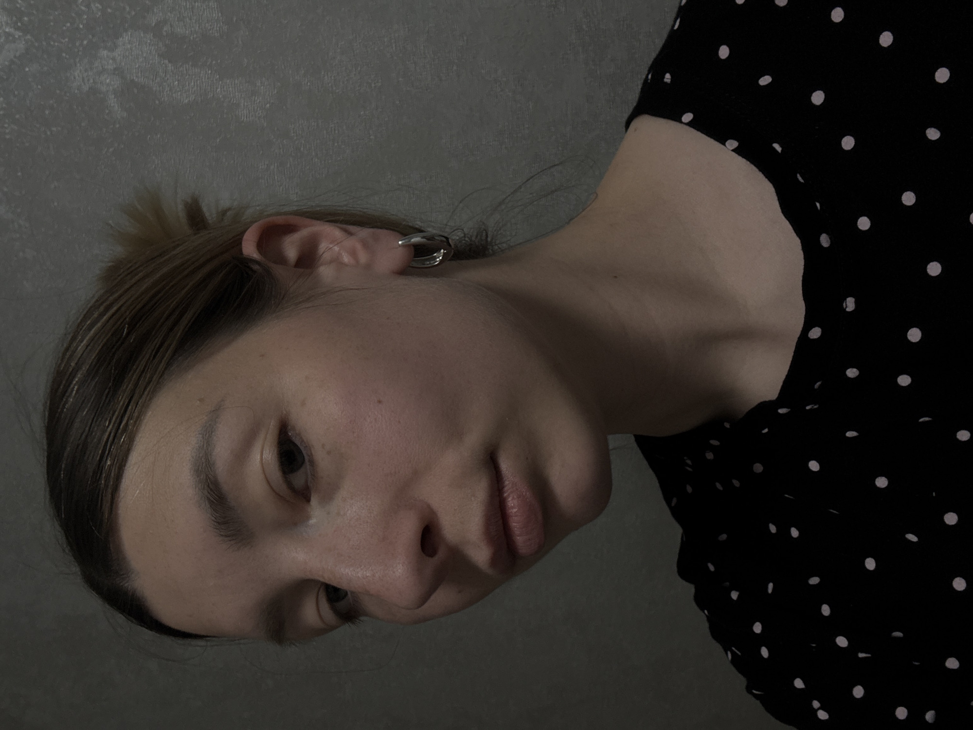 A woman with light skin and brown hair pulled back is shown in a medium shot from the chest up. She is wearing a black blouse with white polka dots. She has a silver hoop earring on her left ear. The background is a grey textured wall. The photograph may have been taken indoors. No location in Moscow, Russia is discernible from the image.