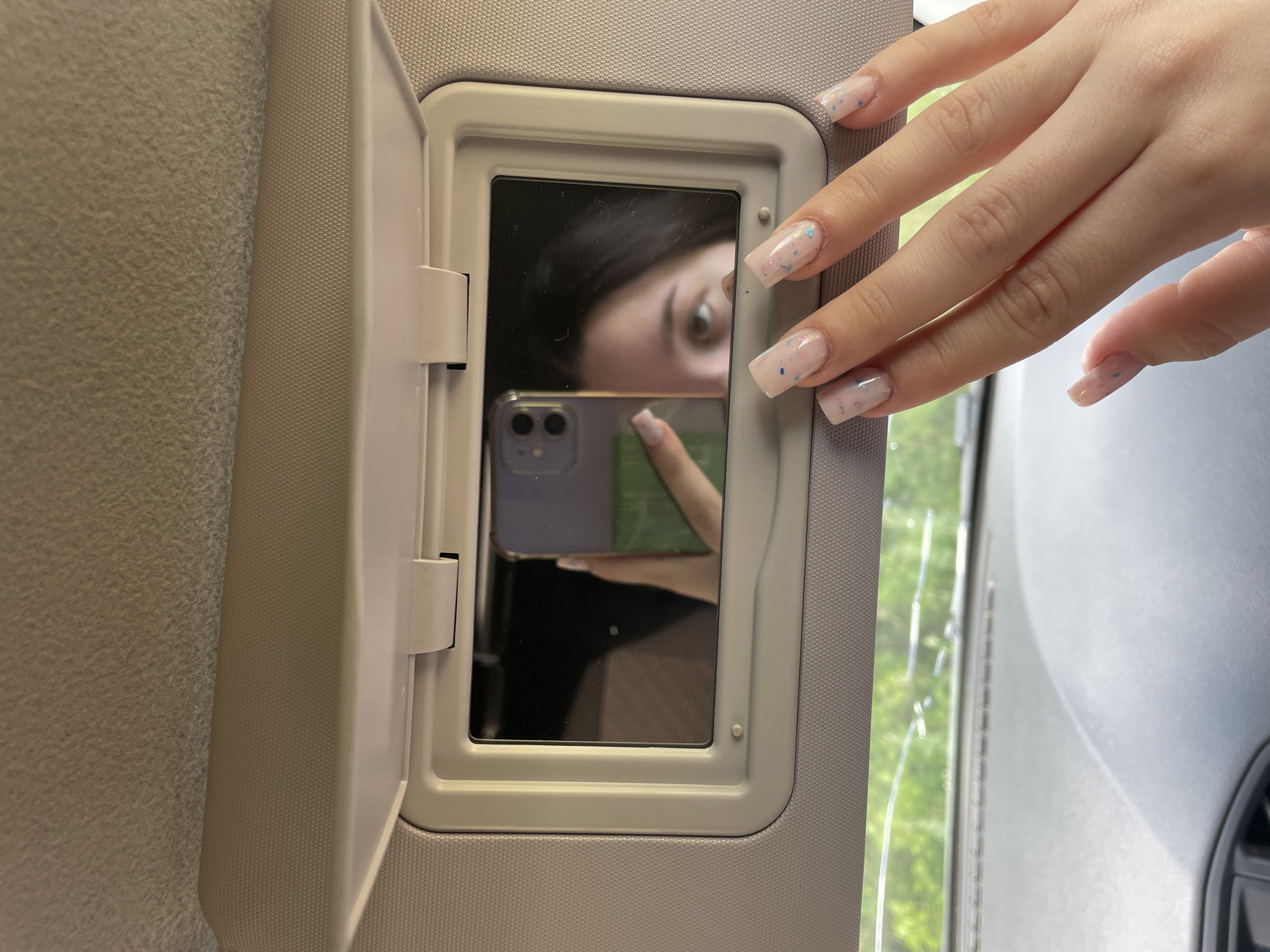 The image shows a selfie taken in a car's visor mirror. A woman's hand with square, light-pink nails adorned with blue and pink speckles is visible in the foreground, touching the mirror. Reflected in the mirror is the woman's face, partially visible, with dark hair and one eye looking towards the camera. She is holding a smartphone, also reflected in the mirror, which is being used to take the picture. The background outside the car appears to be a blurry view of green trees, suggesting an outdoor setting, possibly a rural or suburban area. The interior of the car, including the textured visor and dashboard, is also visible. The location of Nozhovka, Russia is not discernible from the image.