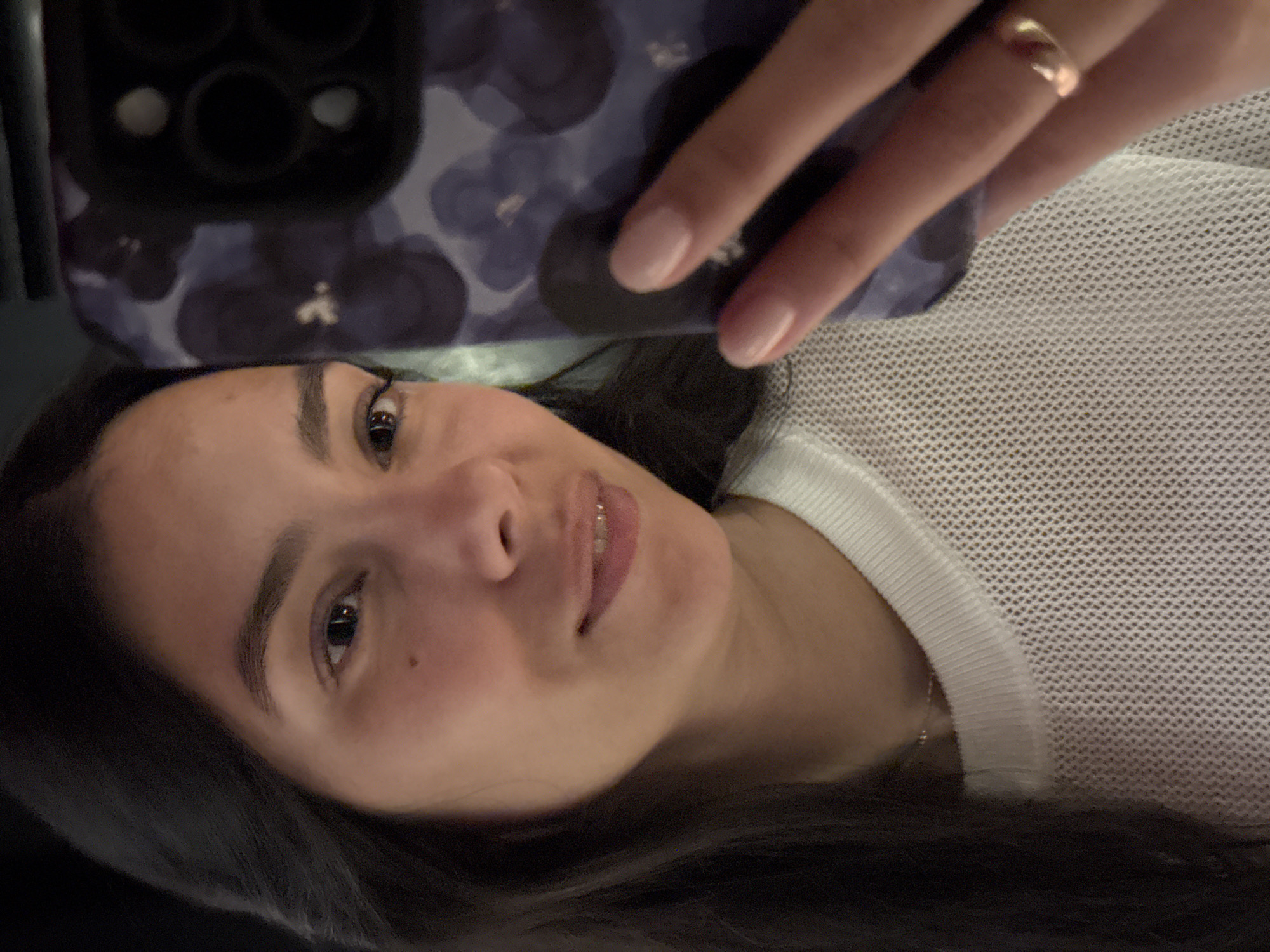 A person with dark hair and light skin is holding a smartphone with a dark-colored case covered in a floral pattern. The person is wearing a white top and a gold ring on their finger. The background is blurry. It is not possible to determine the location or any actions from the provided image.