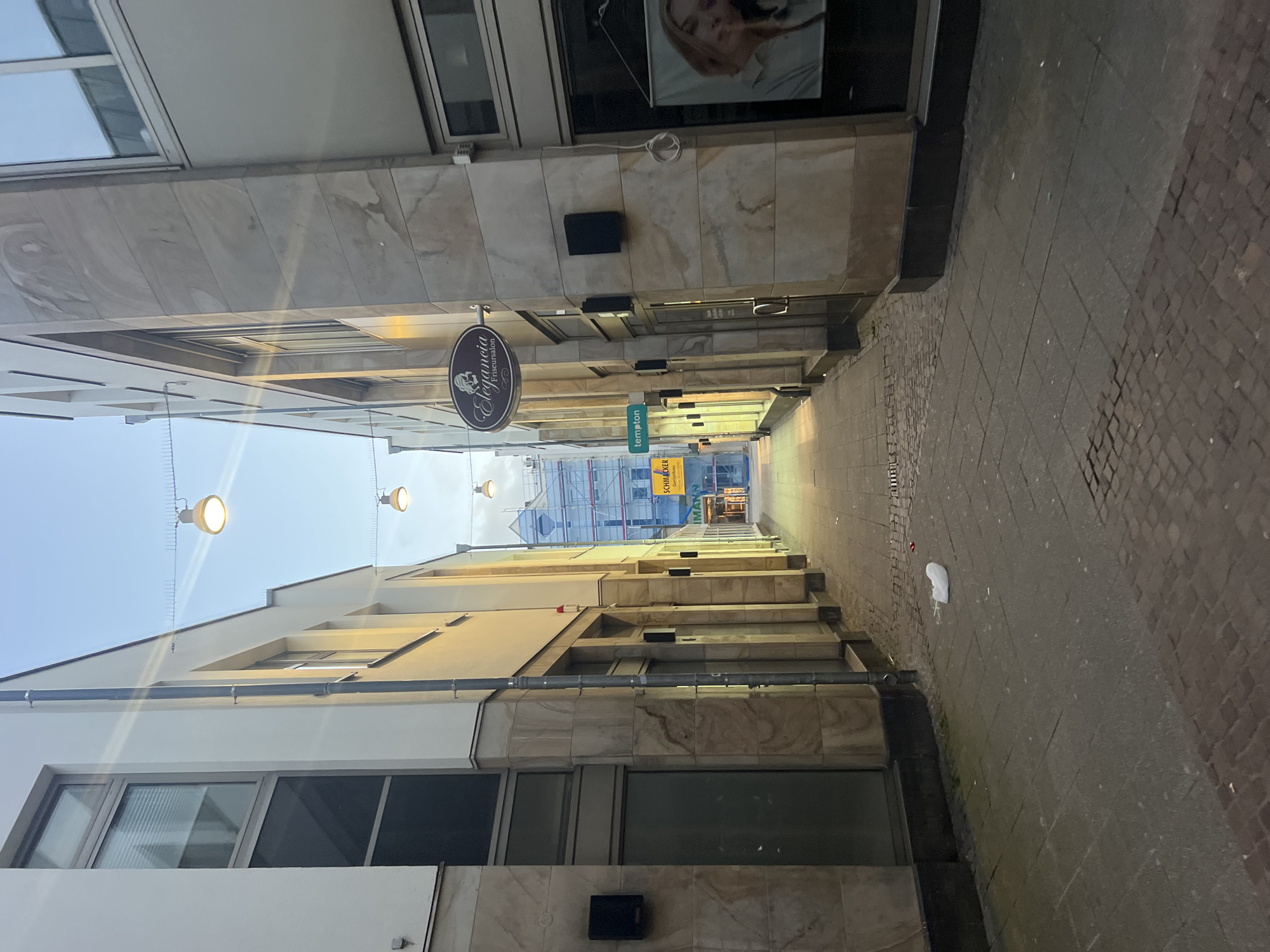 This is a street-level, wide-angle, outdoor photograph of a narrow pedestrian alleyway in the city of Oldenburg, Germany. The alley is flanked by modern, light-colored buildings with stone facades and large windows. Several overhead string lights are visible, some illuminated.
On the left side of the alley, a building features large, dark-tinted windows on the lower level and lighter windows with blinds on the upper levels. A black speaker is mounted to the wall near the lower windows.
On the right side of the alley, a prominent oval sign with the text "Elegancia Friseursalon" (Elegancia Hair Salon) is mounted above the entrance to a business. Below this sign, a black speaker is also mounted. A poster or banner featuring the face of a blonde woman is visible in the window of a shop on the right.
The ground of the alley is paved with cobblestones and rectangular paving stones. A crumpled white plastic bag lies on the ground in the middle of the alleyway, near the center of the frame.
In the background, the alley opens up slightly to reveal a building covered in scaffolding, suggesting ongoing construction or renovation. Signs for "tempton" and "SCHMACKER IMMOBILIEN" are visible further down the alley. The sky above is clear and a pale blue, indicative of twilight. No people are visible in the scene.