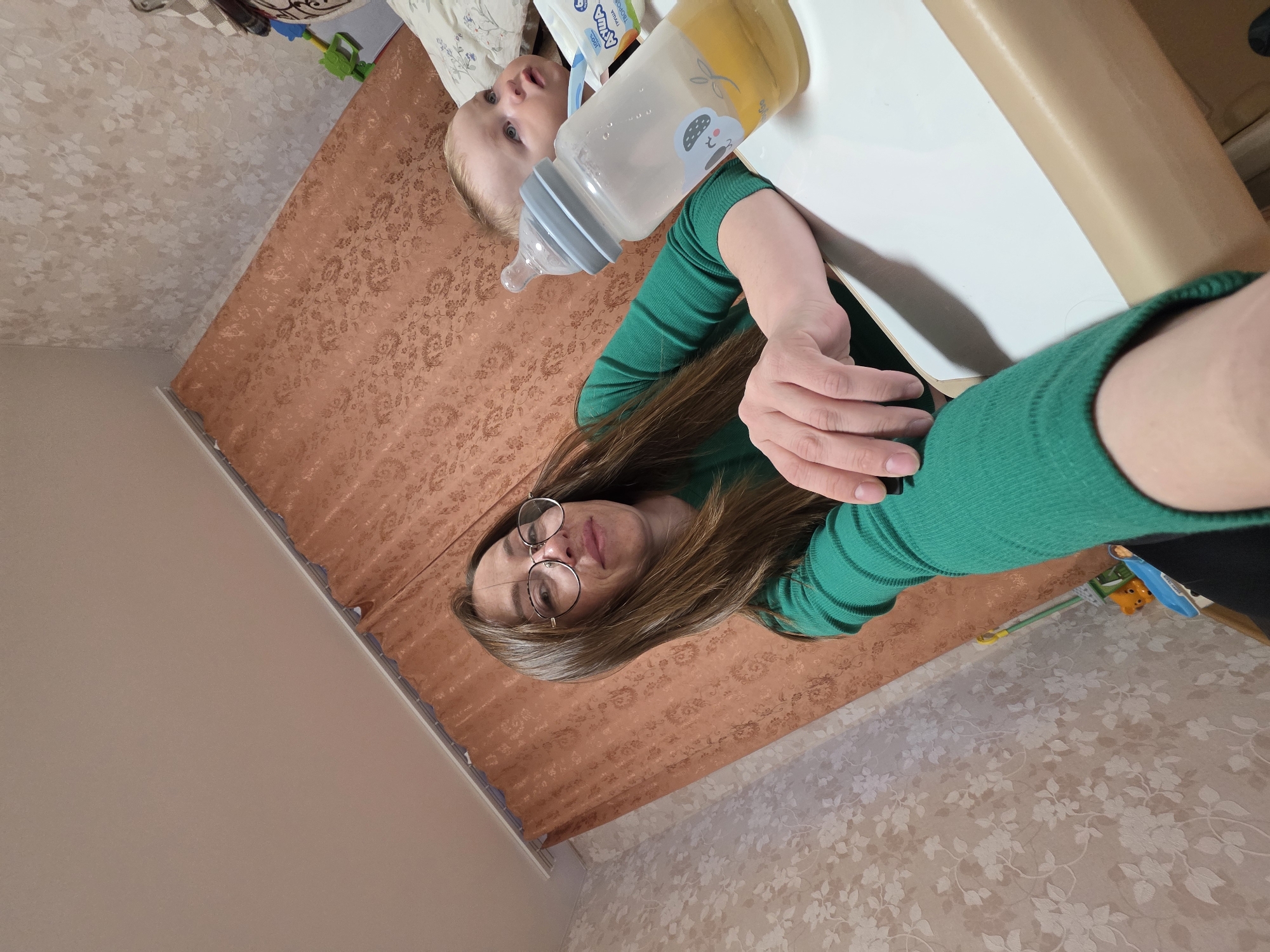 A top-down, skewed perspective photograph captures a woman in a green, long-sleeved shirt holding a baby bottle filled with yellow liquid above a young child lying on a surface. The child, positioned to the left of the frame, gazes upward with wide eyes. The bottle features a light gray lid and a blue koala design on the bottle. The surface the child is on appears to be a cushioned changing pad atop a patterned bedspread. The woman is holding the bottle in a way that the bottle is directly above the baby. The environment suggests an interior setting with patterned wallpaper on the lower-right section of the photo and a solid light-colored wall on the lower left. A white table is on the right side. The location appears to be in a private residence in the city of Unknown, Unknown.