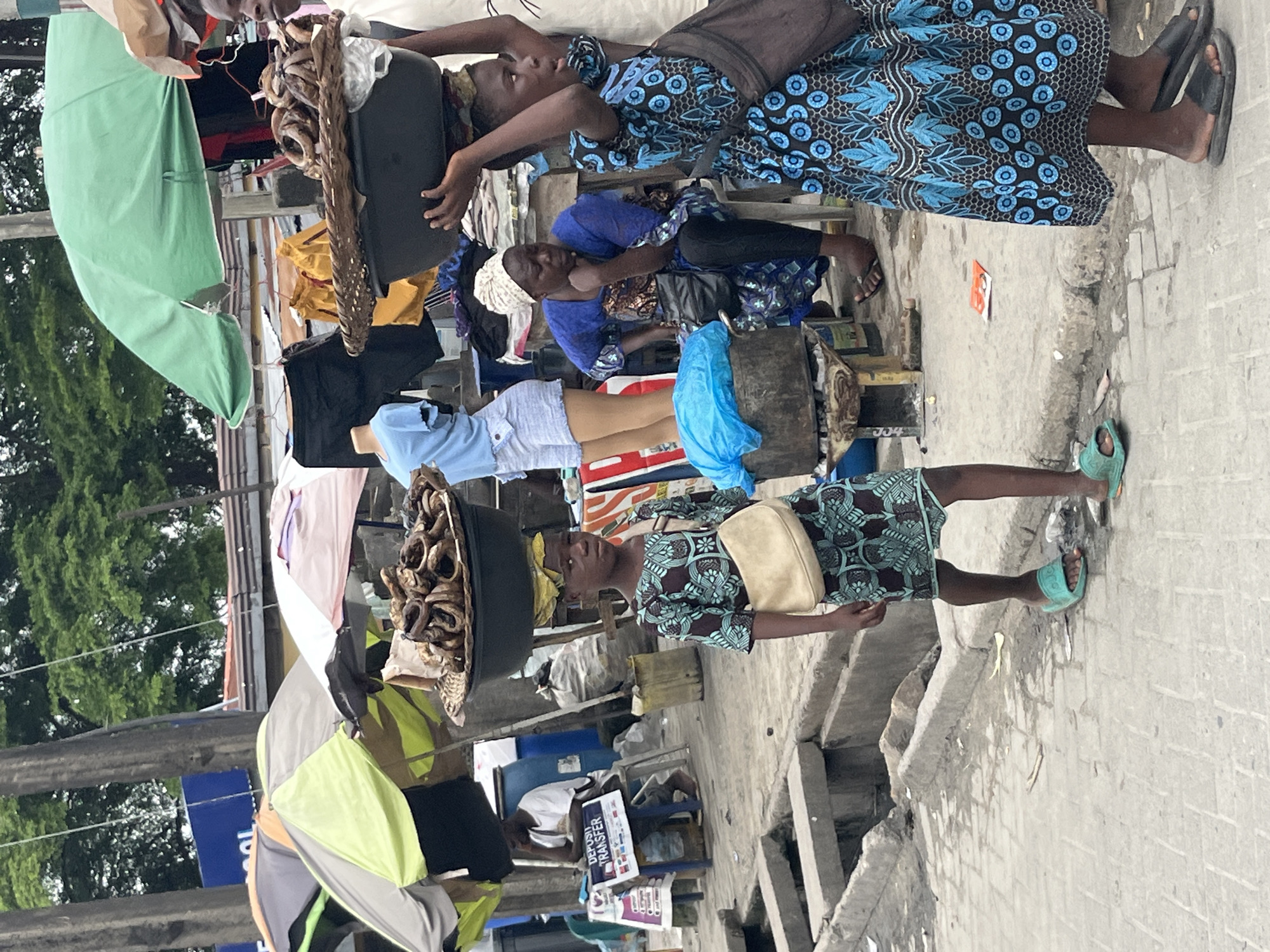 In Ado, Nigeria, a street market bustles with activity under a cloudy sky. A young girl, wearing a green and teal patterned dress and light-colored sandals, carries a large bowl of dried fish on her head. Beside her, a woman in a blue and white patterned dress and headwrap sits, looking down with her chin resting on her hand. Another person, partially visible on the right, wears a blue dress with a black and white circular pattern. Various stalls are set up with umbrellas providing shade, displaying clothing and other goods. In the background, a man sits at a table with a sign that reads "DEPOSIT TRANSFER." The ground is paved with concrete and bricks, and a drainage channel runs along the side of the street. The overall scene depicts a vibrant marketplace, full of commerce and daily life.