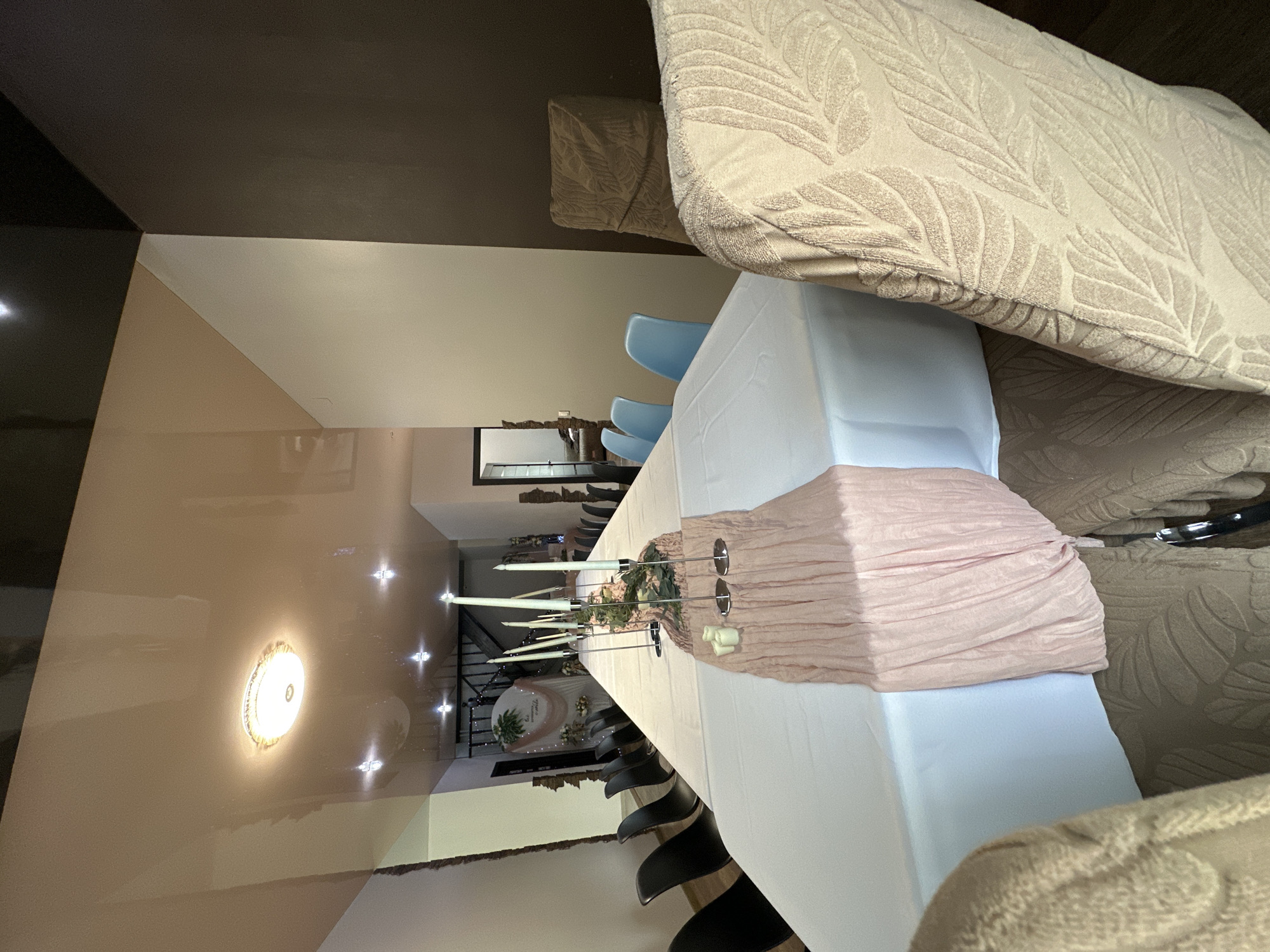 Here is a forensic description of the image:
The image shows an interior setting, likely a banquet or dining room in Unknown, Unknown. The focal point is a long table draped with a white tablecloth. Atop the tablecloth, a pale pink fabric is draped, seemingly bunched or gathered in places to create texture. Centerpieces consisting of tall, white candles in metallic holders are placed along the table, interspersed with floral arrangements containing green foliage.
Chairs are arranged around the table. Light blue chairs are visible at one end, with a row of dark, possibly black chairs extending along the side of the table.
In the background, there is a view down the length of the room, with a glimpse of a door/opening and further arrangements including what appears to be a circular floral decoration. The ceiling has a light fixture, and the walls have a dual-tone paint scheme, with areas of light and darker shades.
In the foreground, parts of upholstered furniture with a textured, leaf-like pattern in beige are visible, suggesting that the image was taken from the perspective of someone seated or standing near these furnishings.